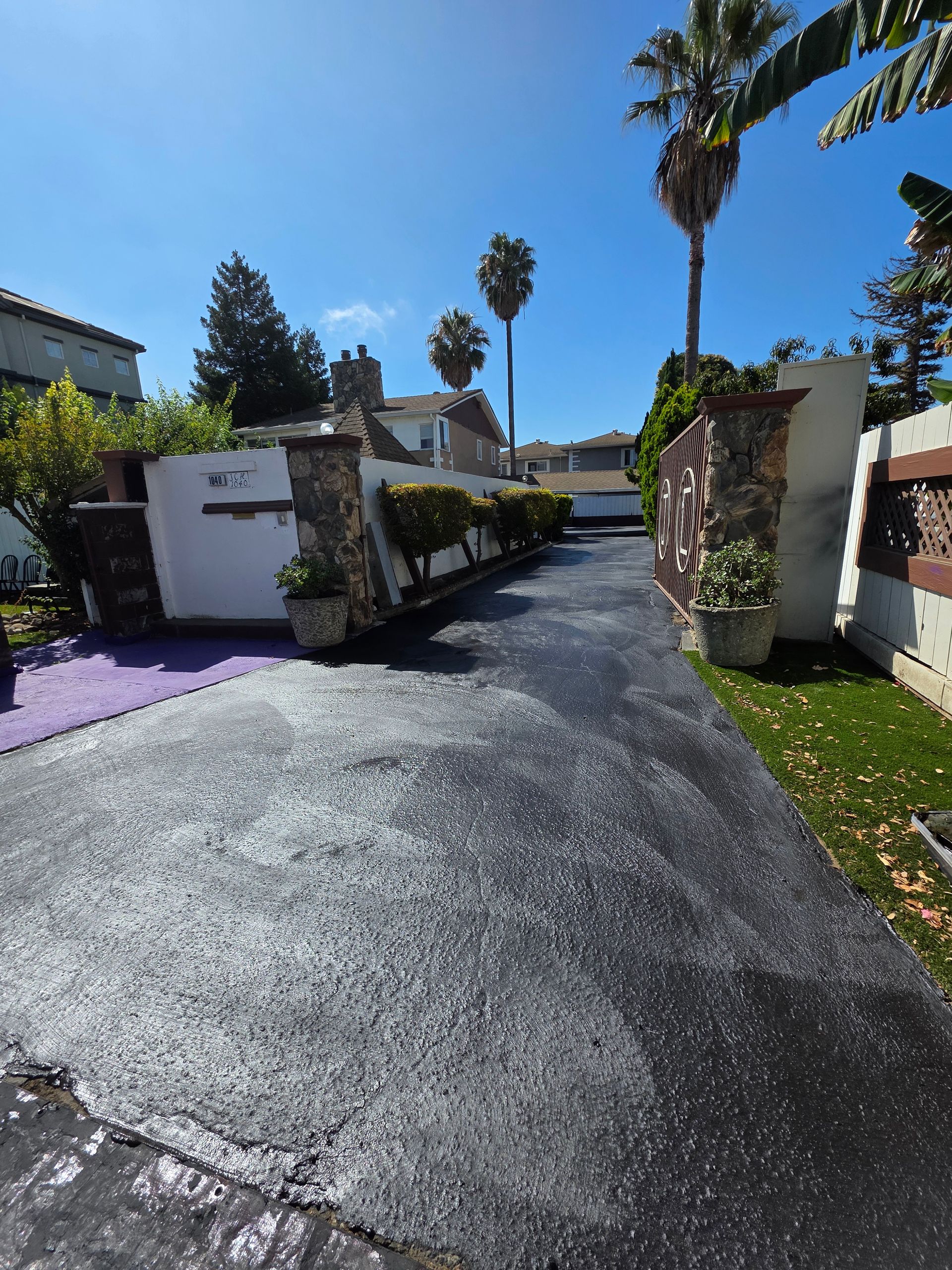 A freshly paved black driveway leads between two stone pillars and white walls, with palm trees and a clear blue sky above.
