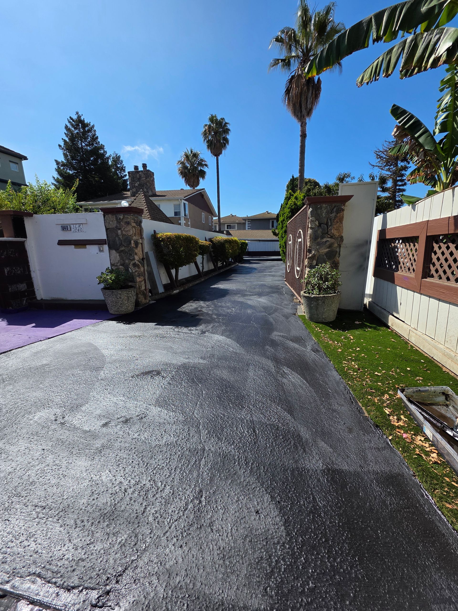 A driveway with fresh black asphalt, framed by stone pillars, white walls, and palm trees under a bright blue sky.