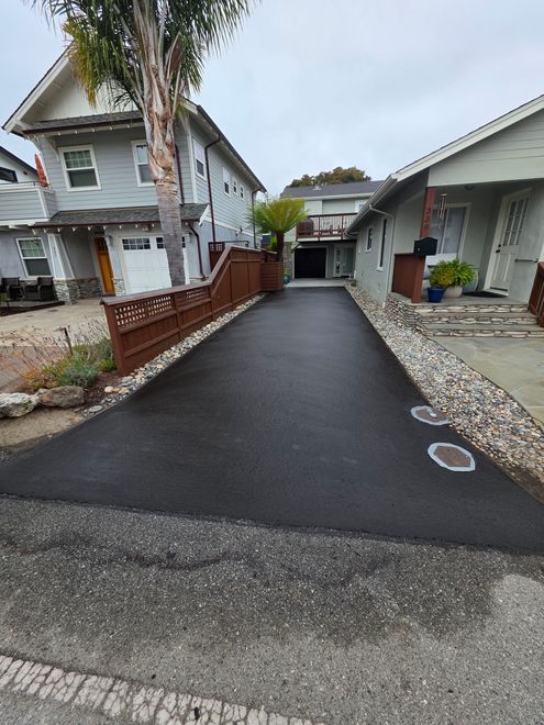 A newly paved black asphalt driveway between two houses, bordered by light-colored decorative rocks and a wooden fence.