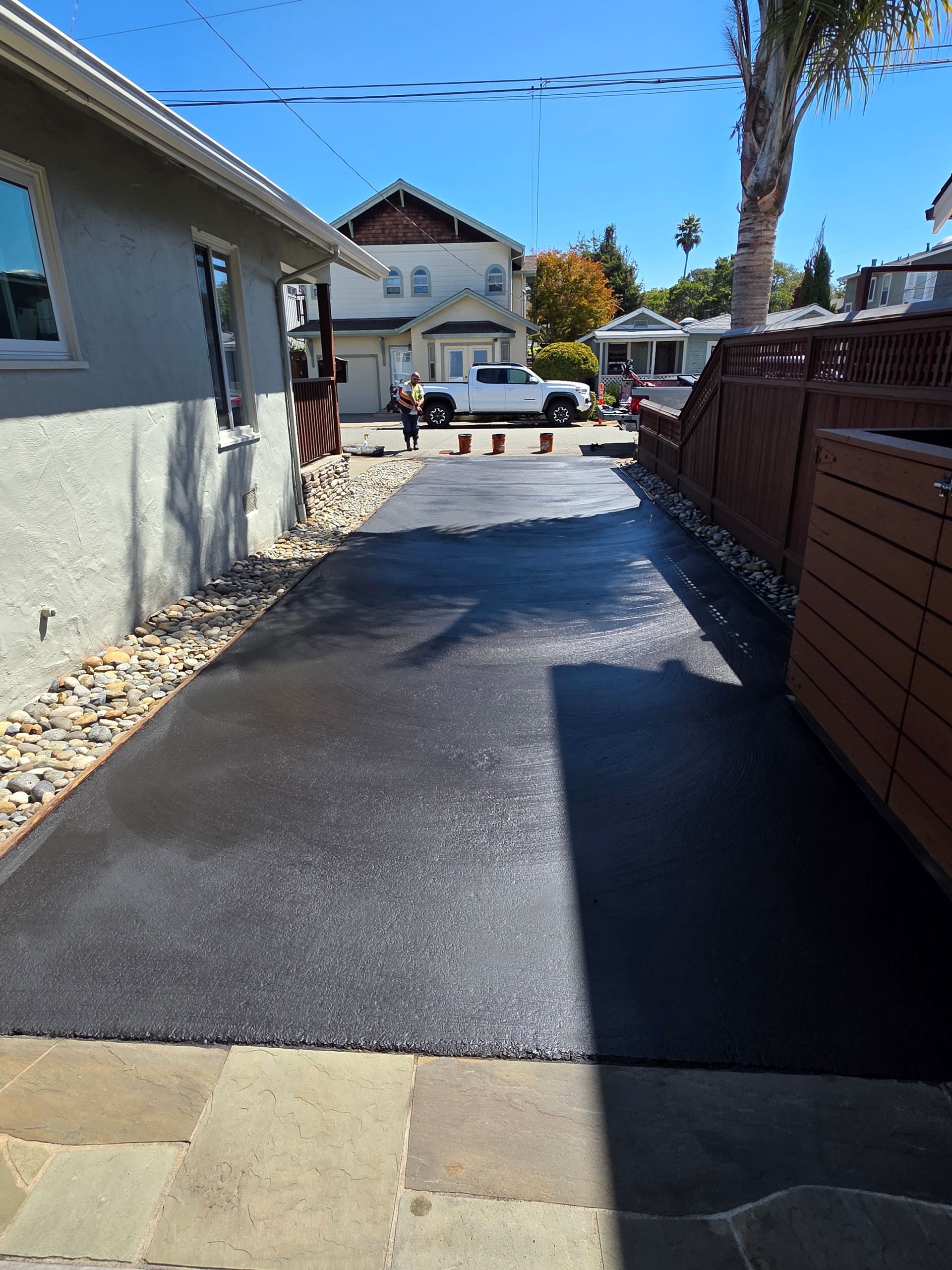 A newly paved asphalt driveway runs between a house and a wooden fence, leading toward a street with parked vehicles.