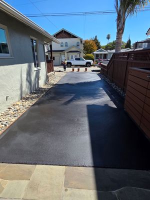 A newly paved, dark asphalt driveway extends between a light grey house and a brown wooden fence on a sunny day.