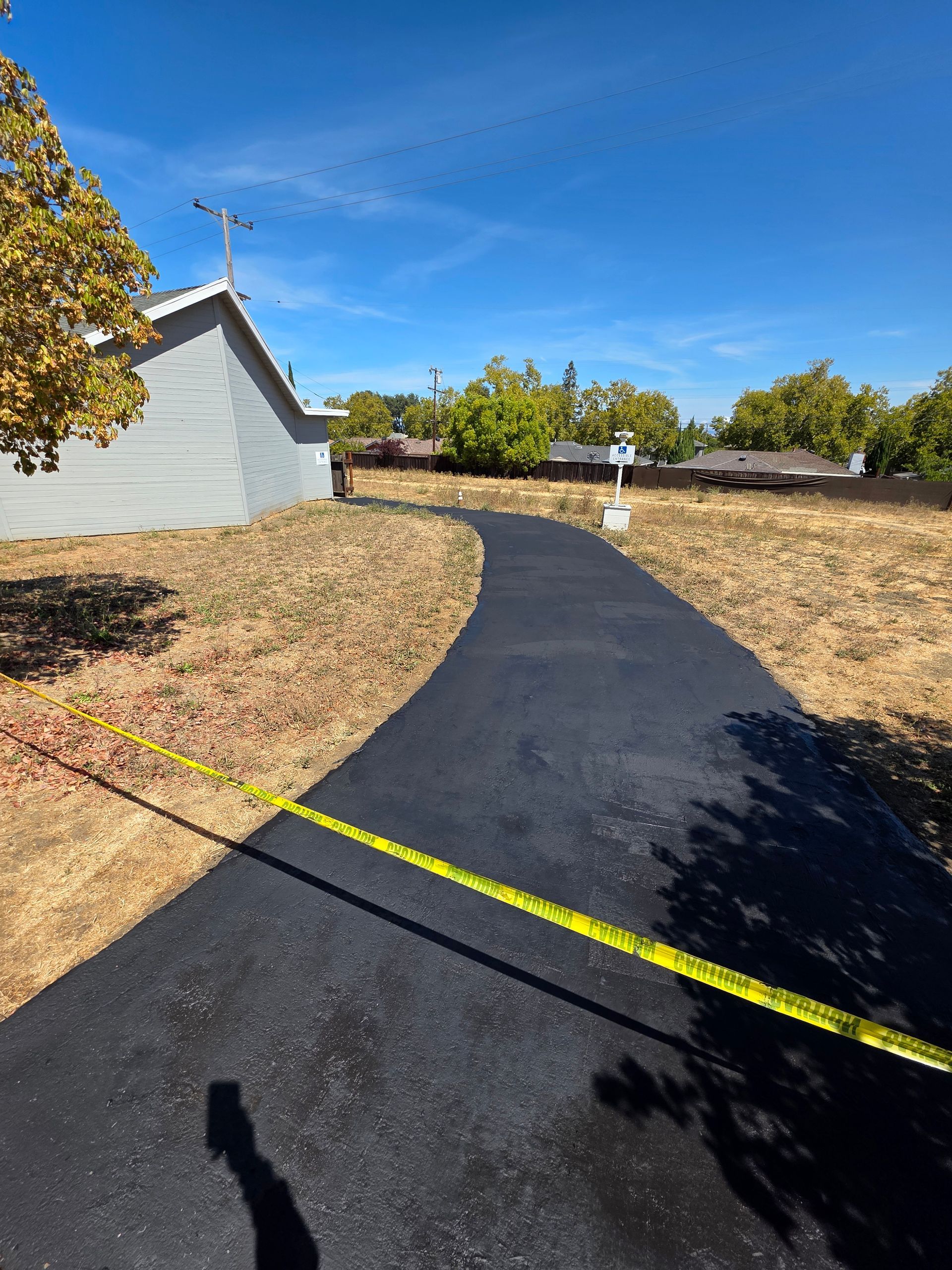 A freshly paved asphalt path leads around the side of a light gray house, blocked by yellow caution tape.