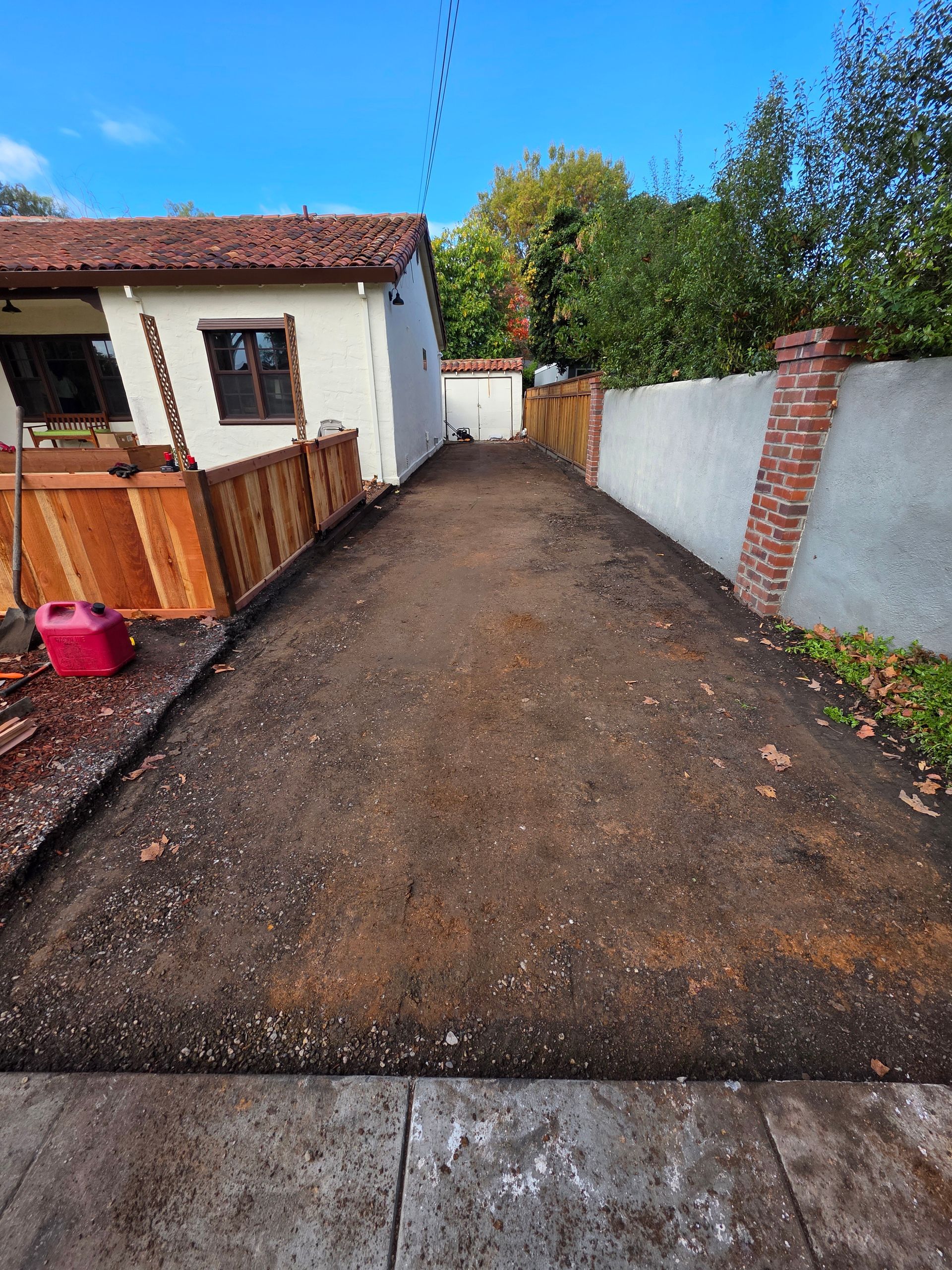 A view of a freshly cleared dirt driveway next to a white house, a wooden fence, and a brick-accented stucco wall.