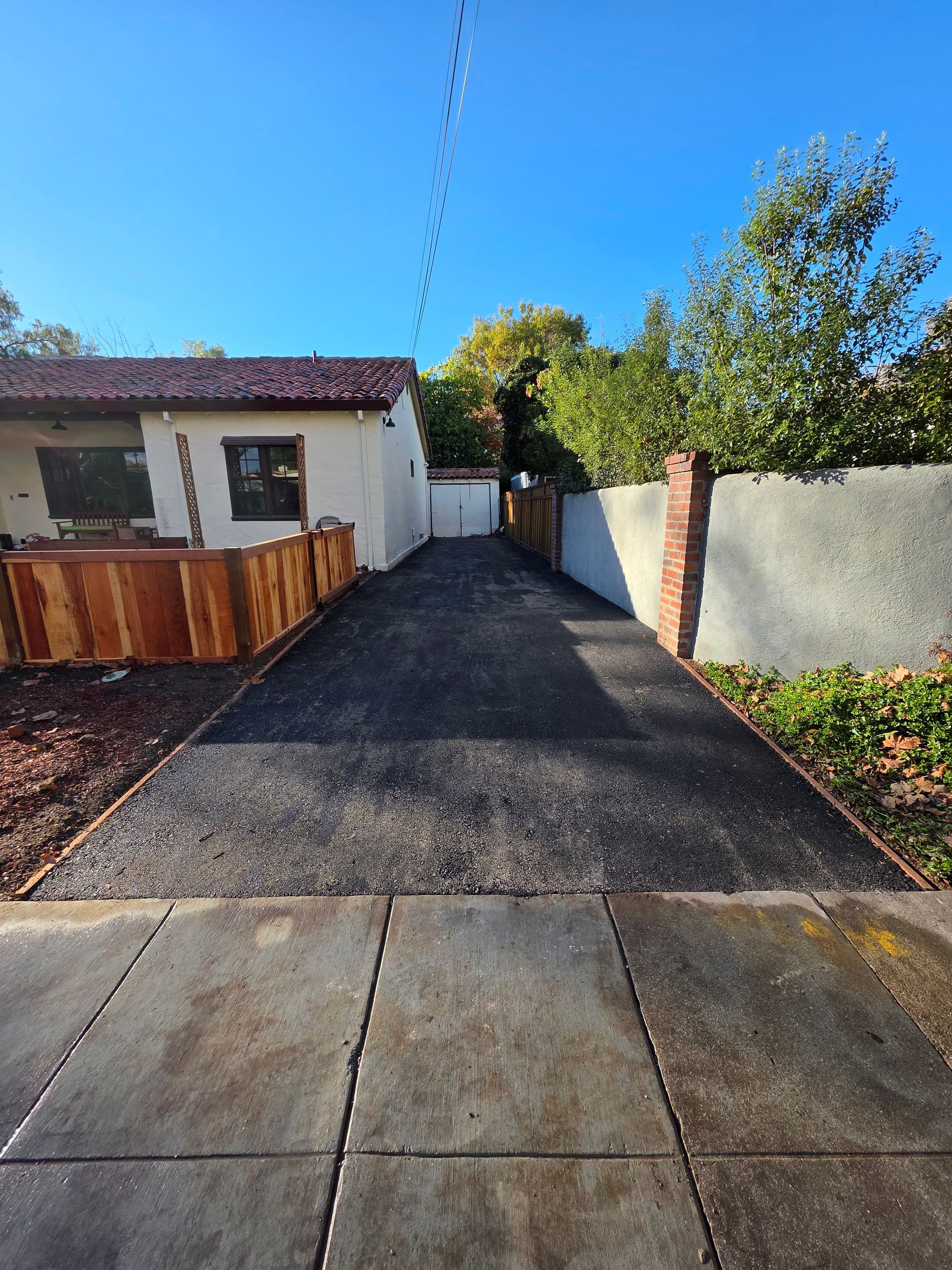 A new gravel driveway leads from a concrete sidewalk toward a house with a wooden fence and a white wall under a blue sky.