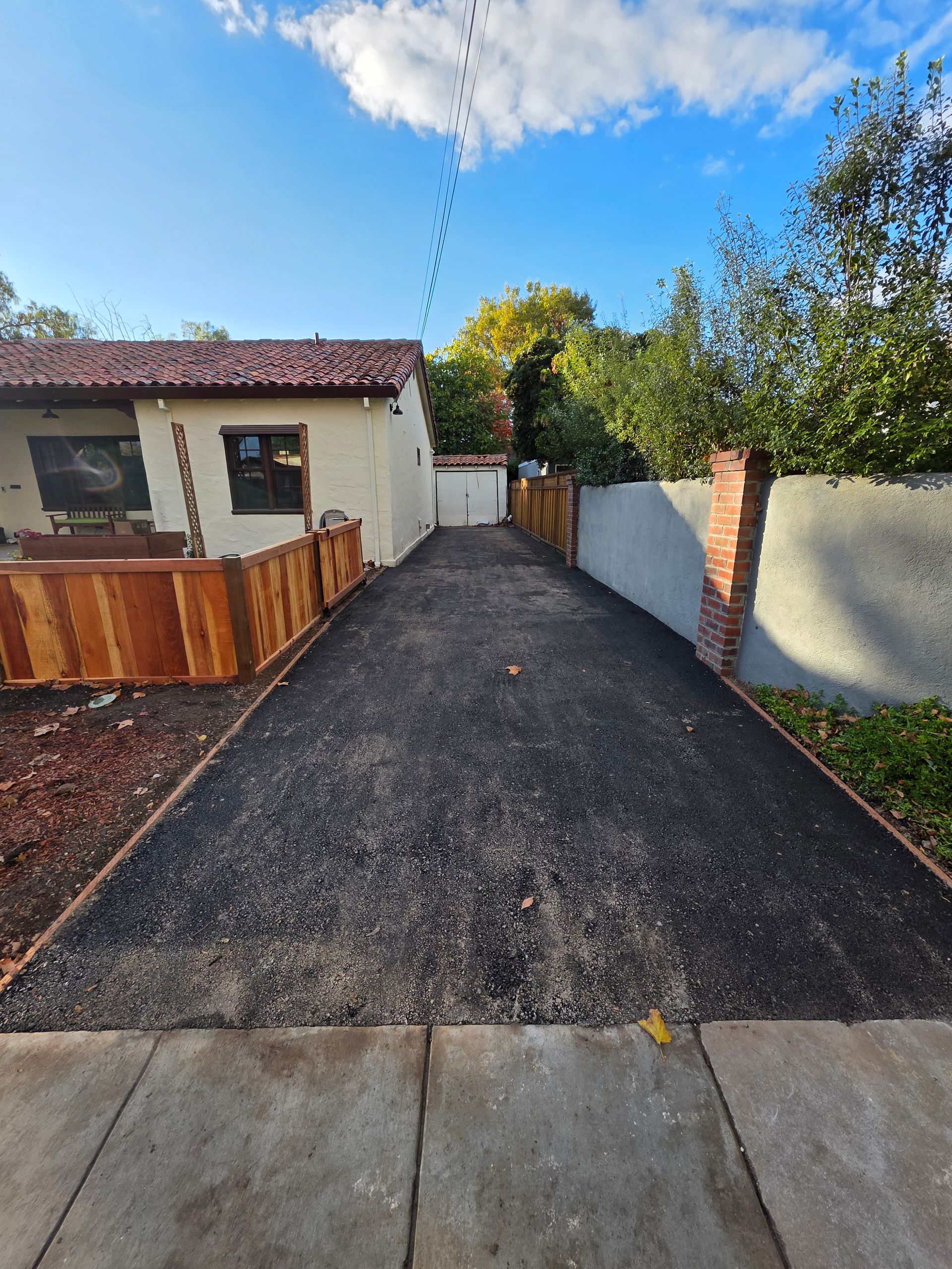 A newly paved dark asphalt driveway leads between a residential house with a wooden fence and a wall to a rear garage.