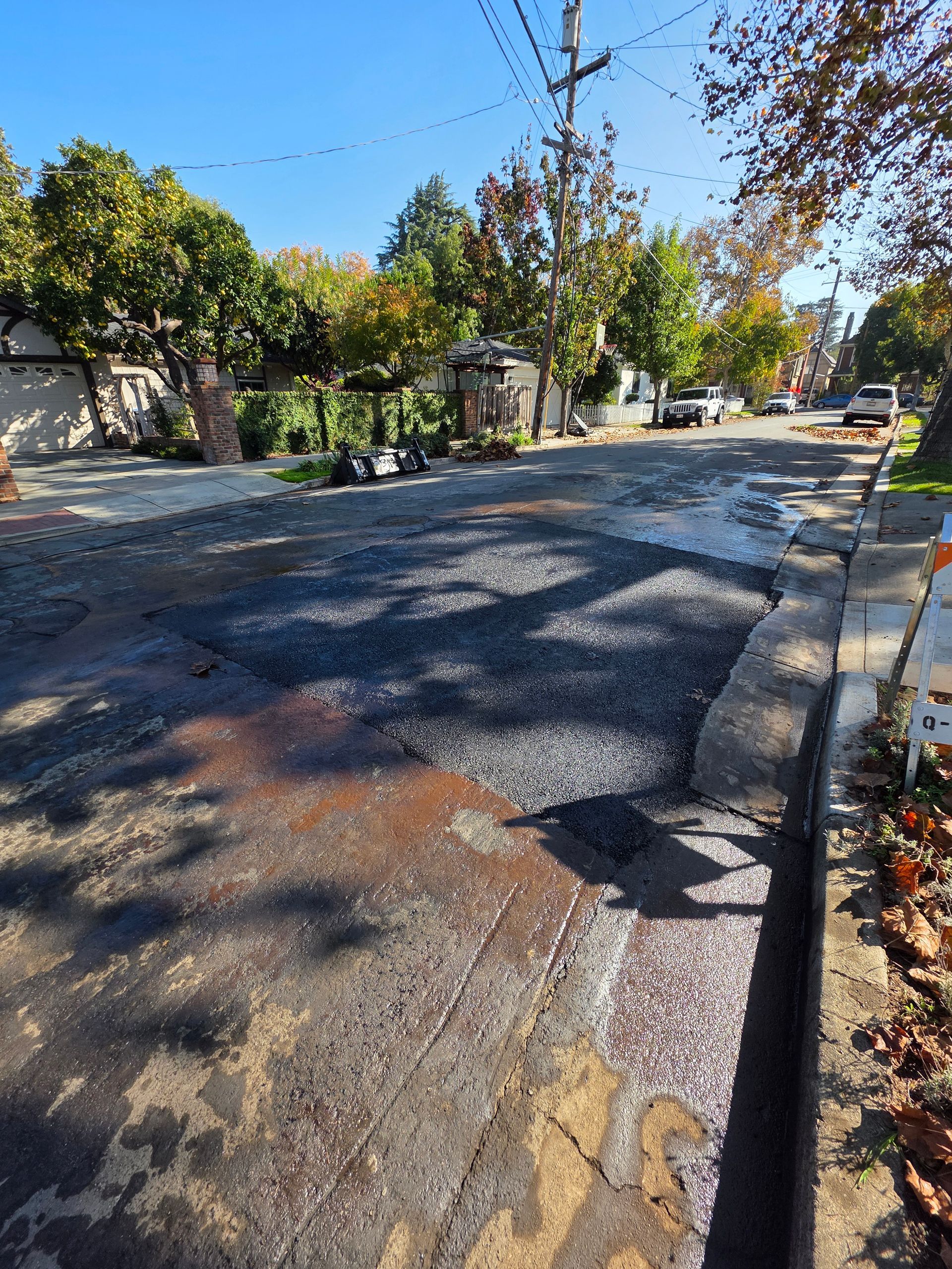 A residential street with a recently patched asphalt section, light-colored gravel, and surrounding autumn trees.