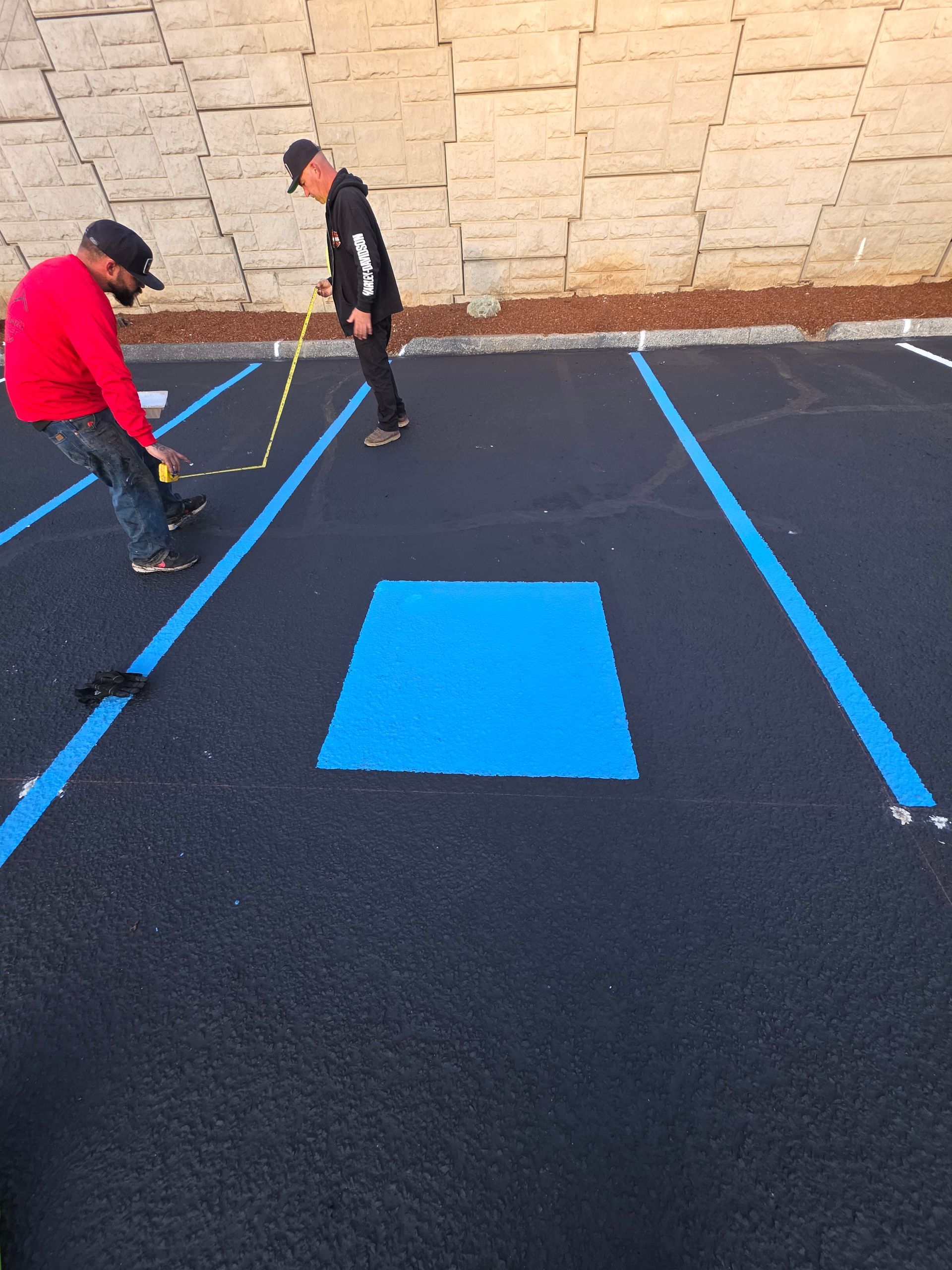 Two people measure and mark a newly painted blue handicap parking space in a parking lot.
