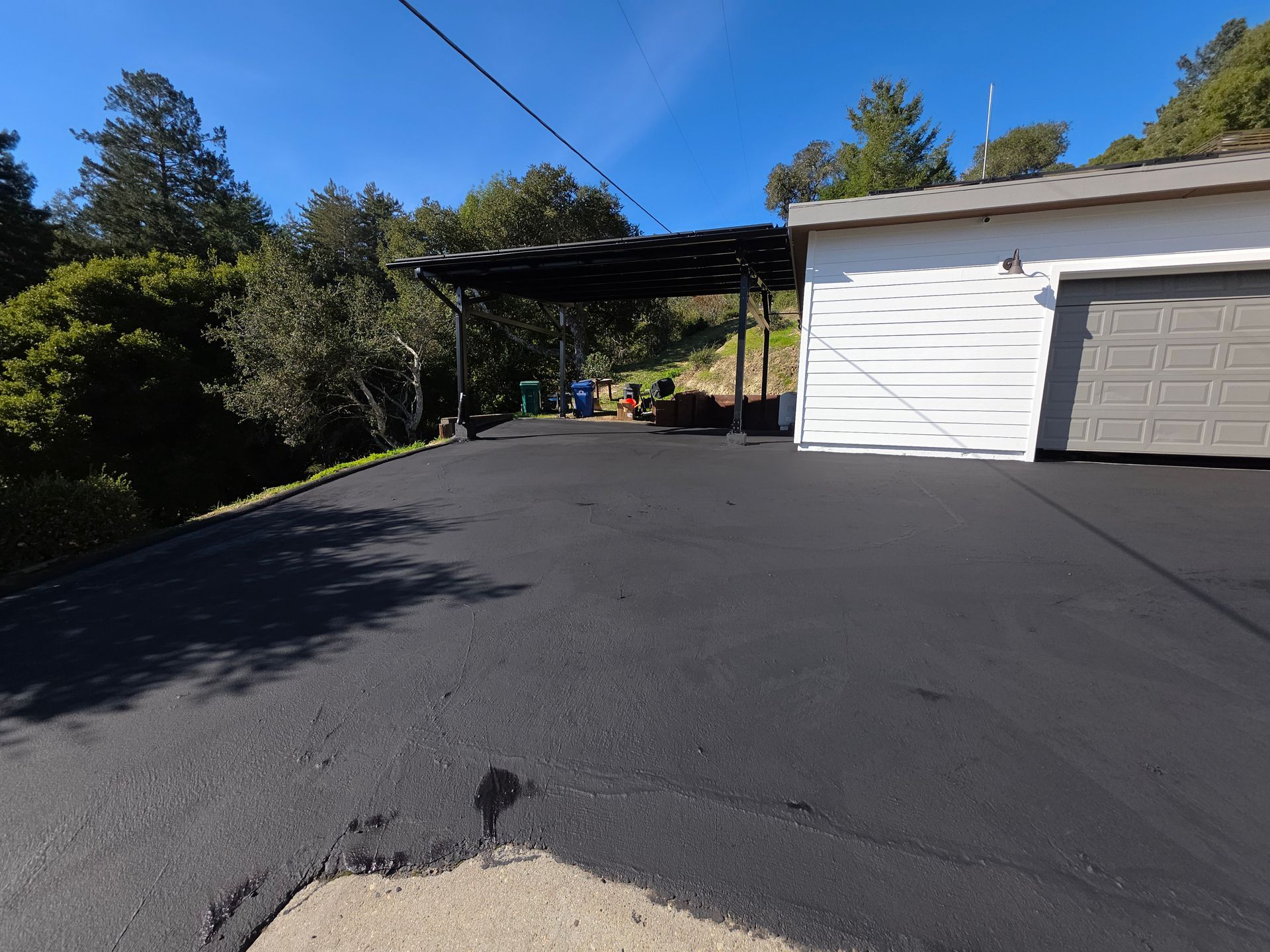 A newly paved black asphalt driveway leading to a white garage and a carport in a wooded area under a blue sky.