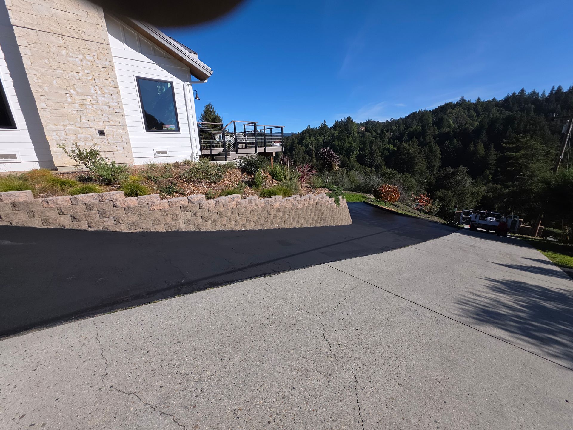 A split driveway featuring a weathered concrete section and a new black asphalt section, leading to a house with stonework.