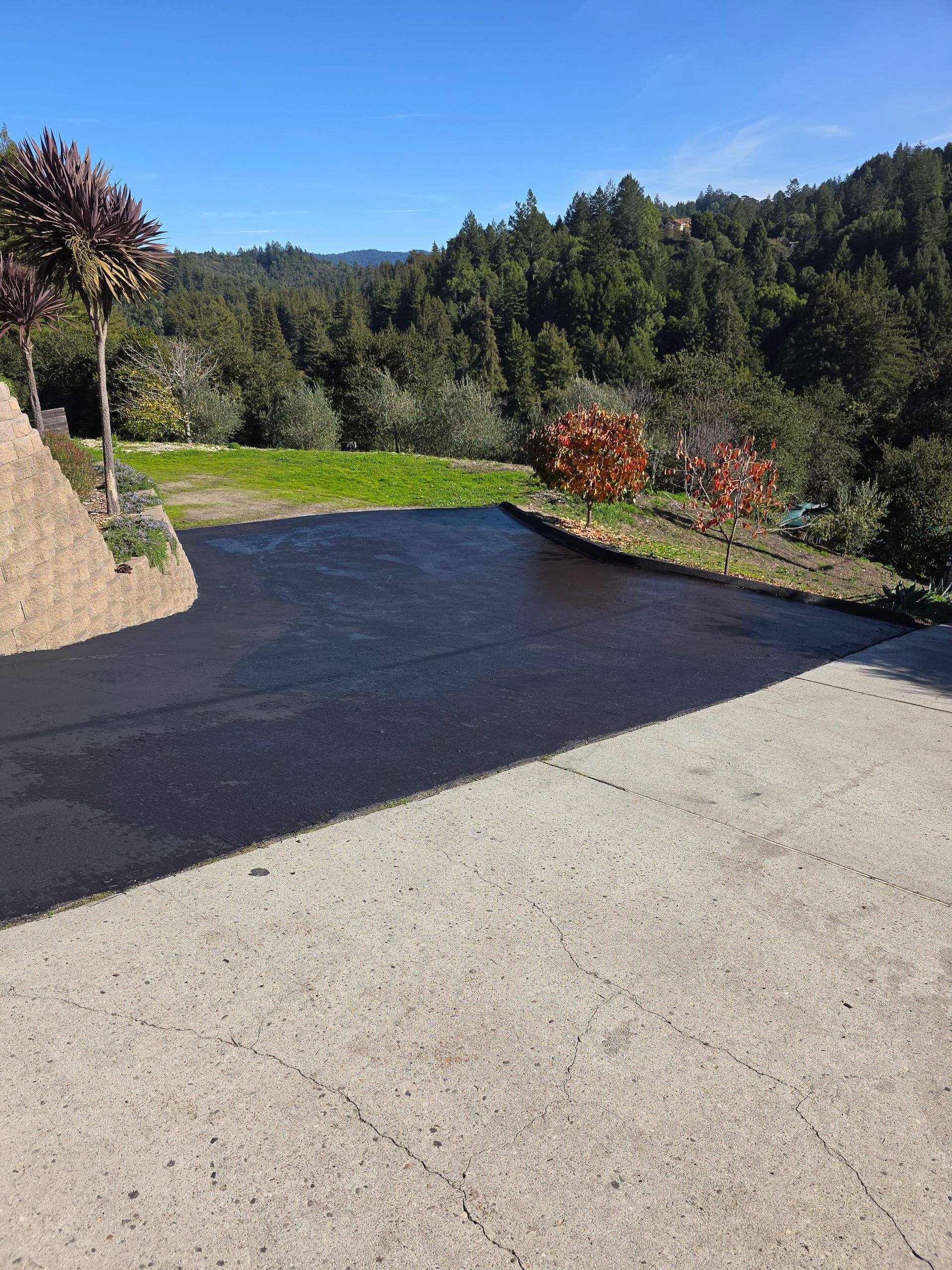 A paved driveway transitions from light-colored concrete to fresh, dark asphalt, overlooking a forest under a blue sky.