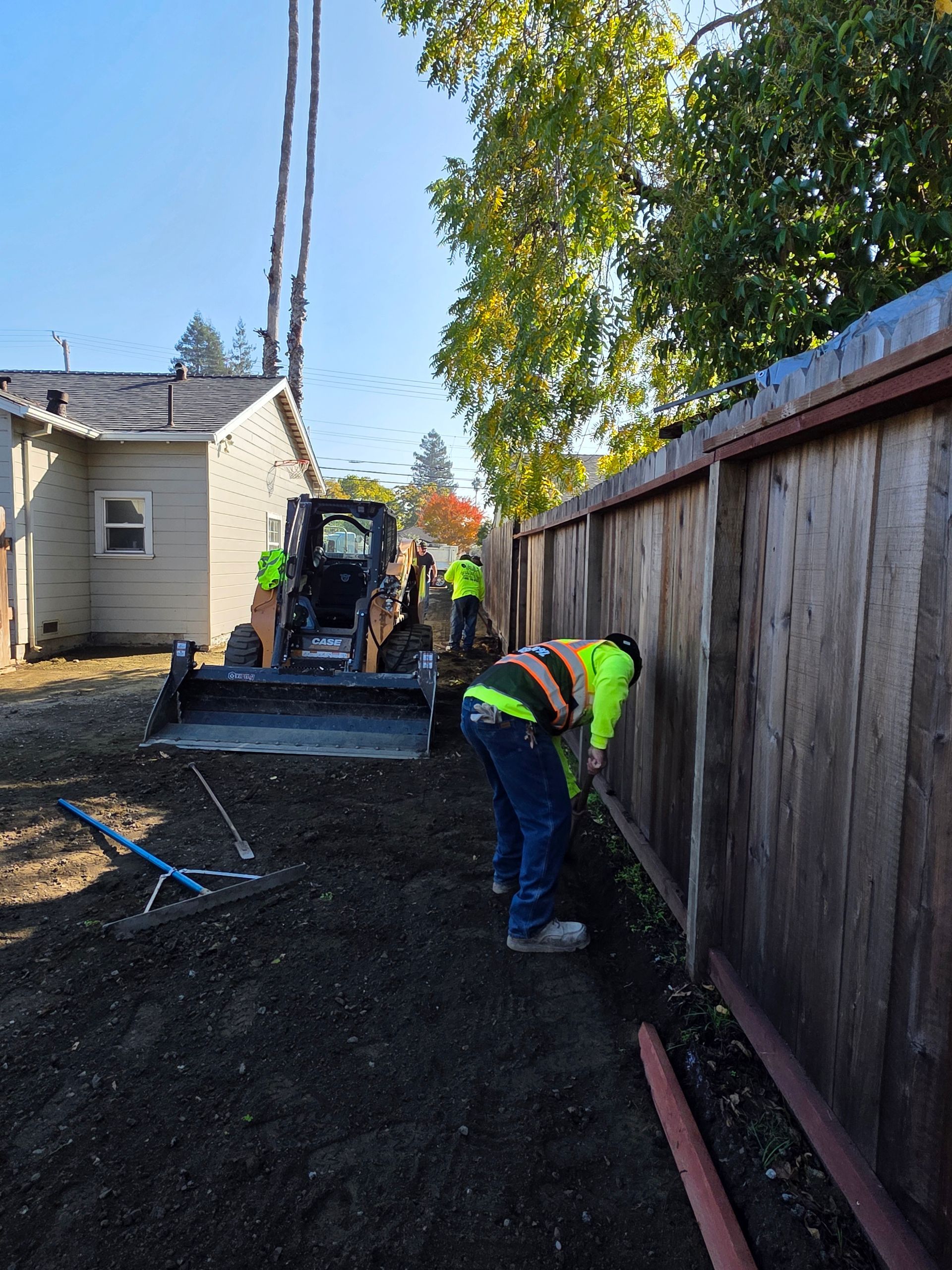 Workers in high-visibility vests work alongside a wooden fence next to a house and a skid steer loader.