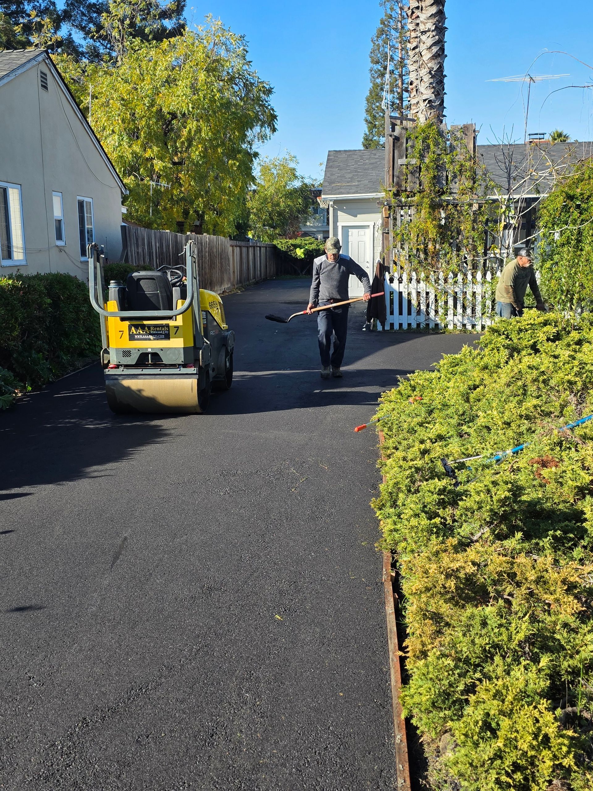 Two workers operate a road roller on a freshly paved residential driveway on a sunny day.