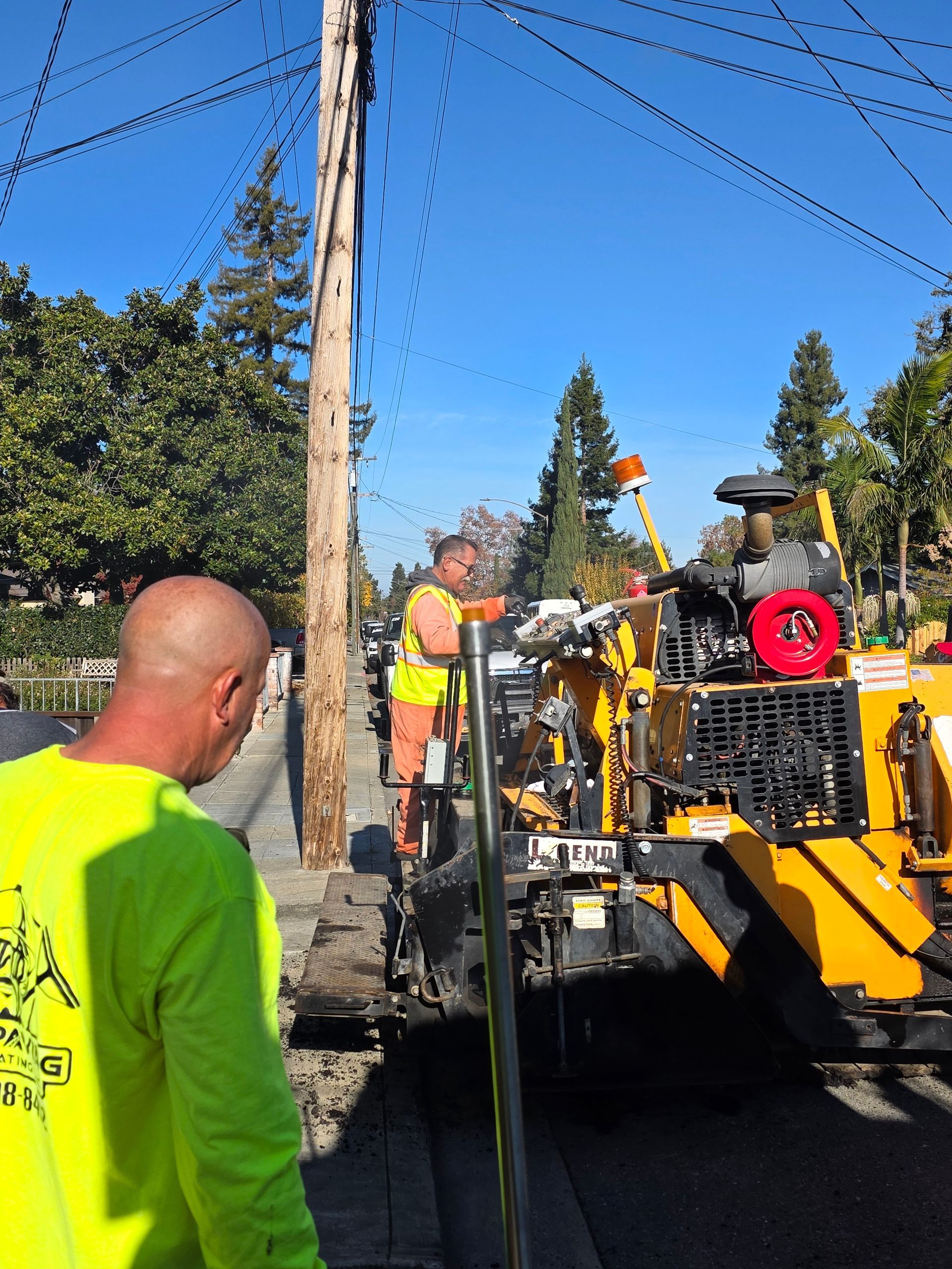 Two workers in high-visibility neon gear operate heavy paving machinery on a sunny, tree-lined residential street.