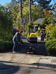 A worker uses a plate compactor on a driveway while another drives a small yellow steamroller in a residential garden.