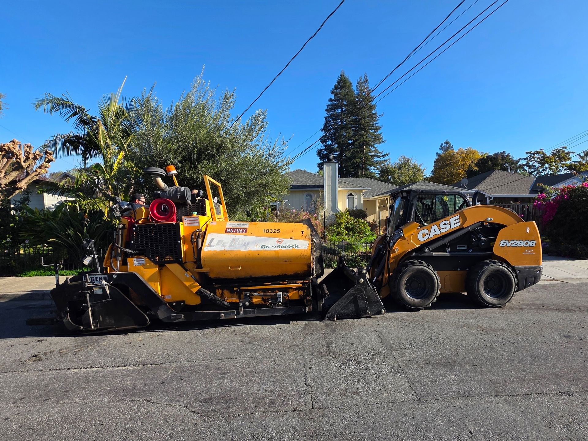 A yellow road paving machine and a Case skid-steer loader parked on an asphalt residential street.