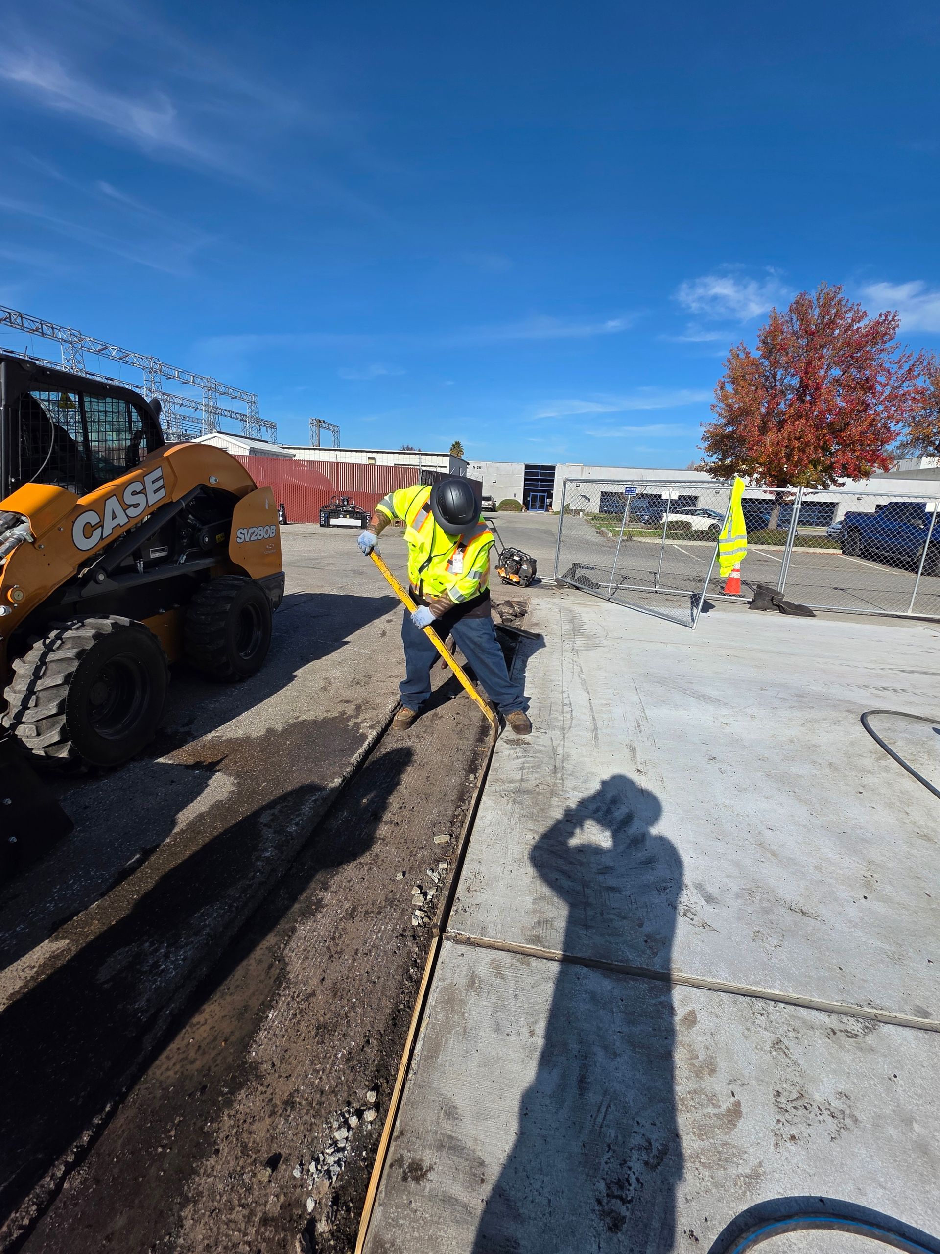 A construction worker in a high-visibility yellow vest uses a tool to clear dirt along a concrete edge near a skid steer.