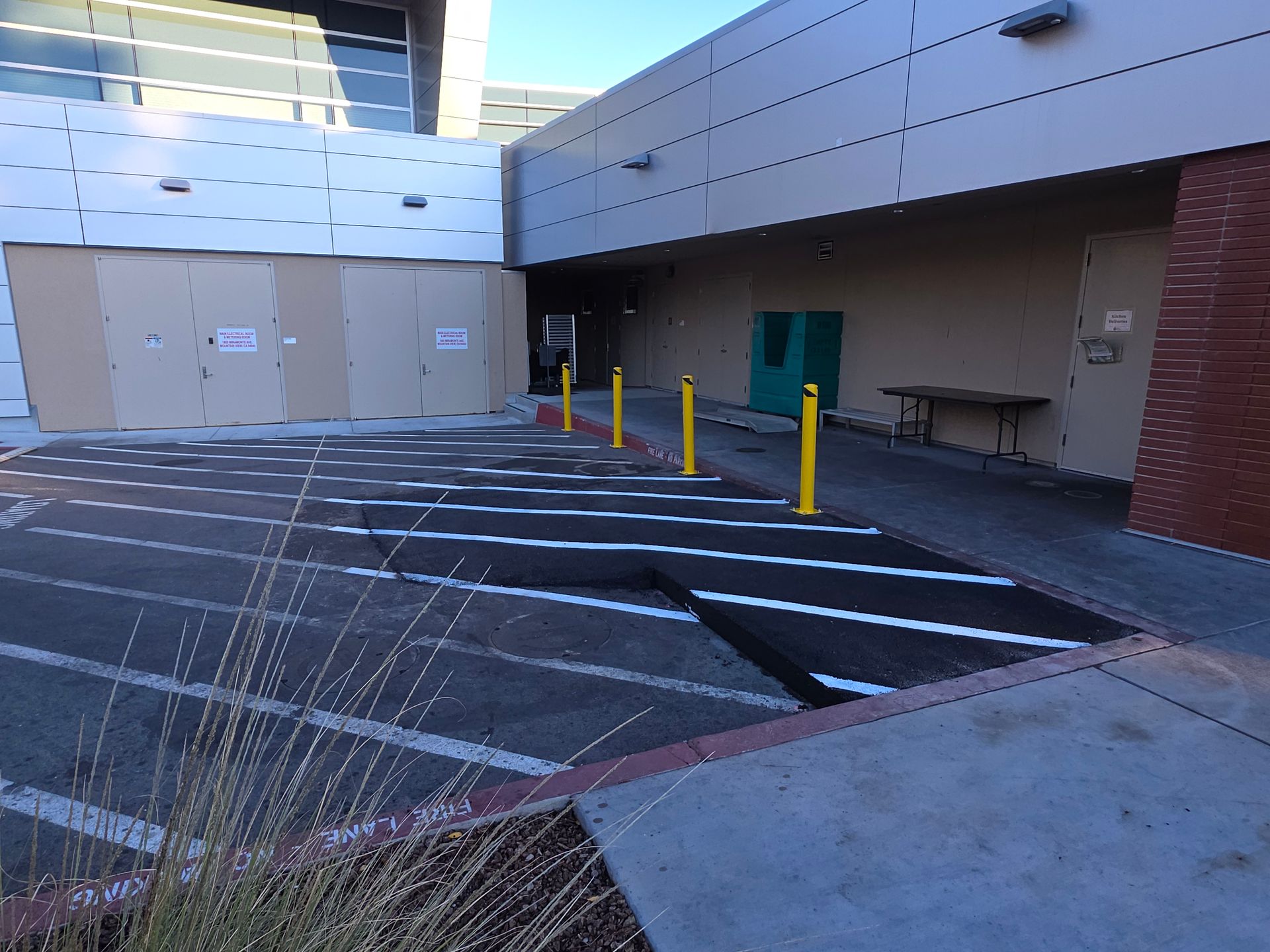 An outdoor paved area with diagonal parking stripes, yellow bollards, and a building entrance featuring beige panels.