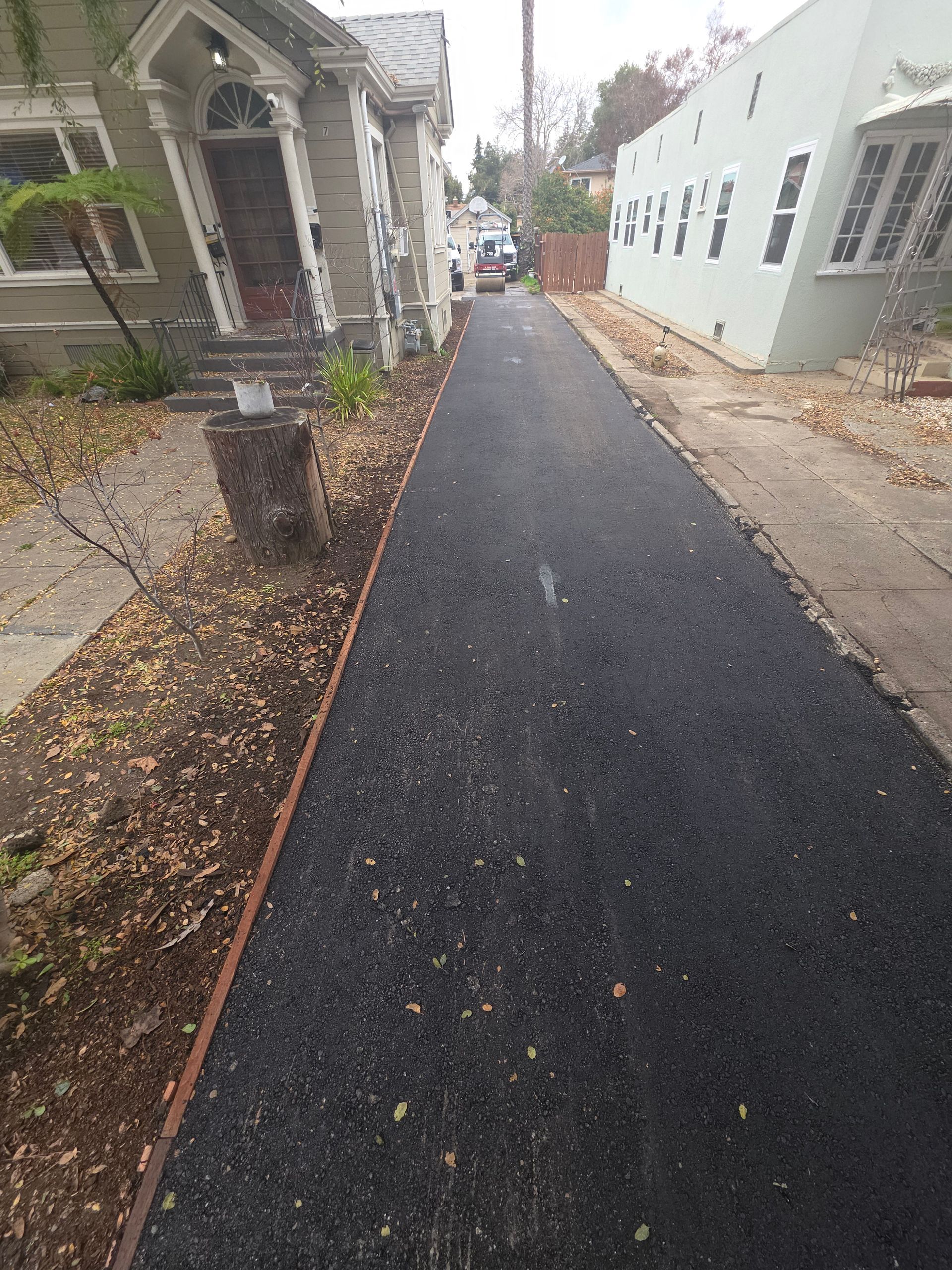 A newly paved, dark asphalt driveway runs between two residential houses with houses and fall leaves on both sides.