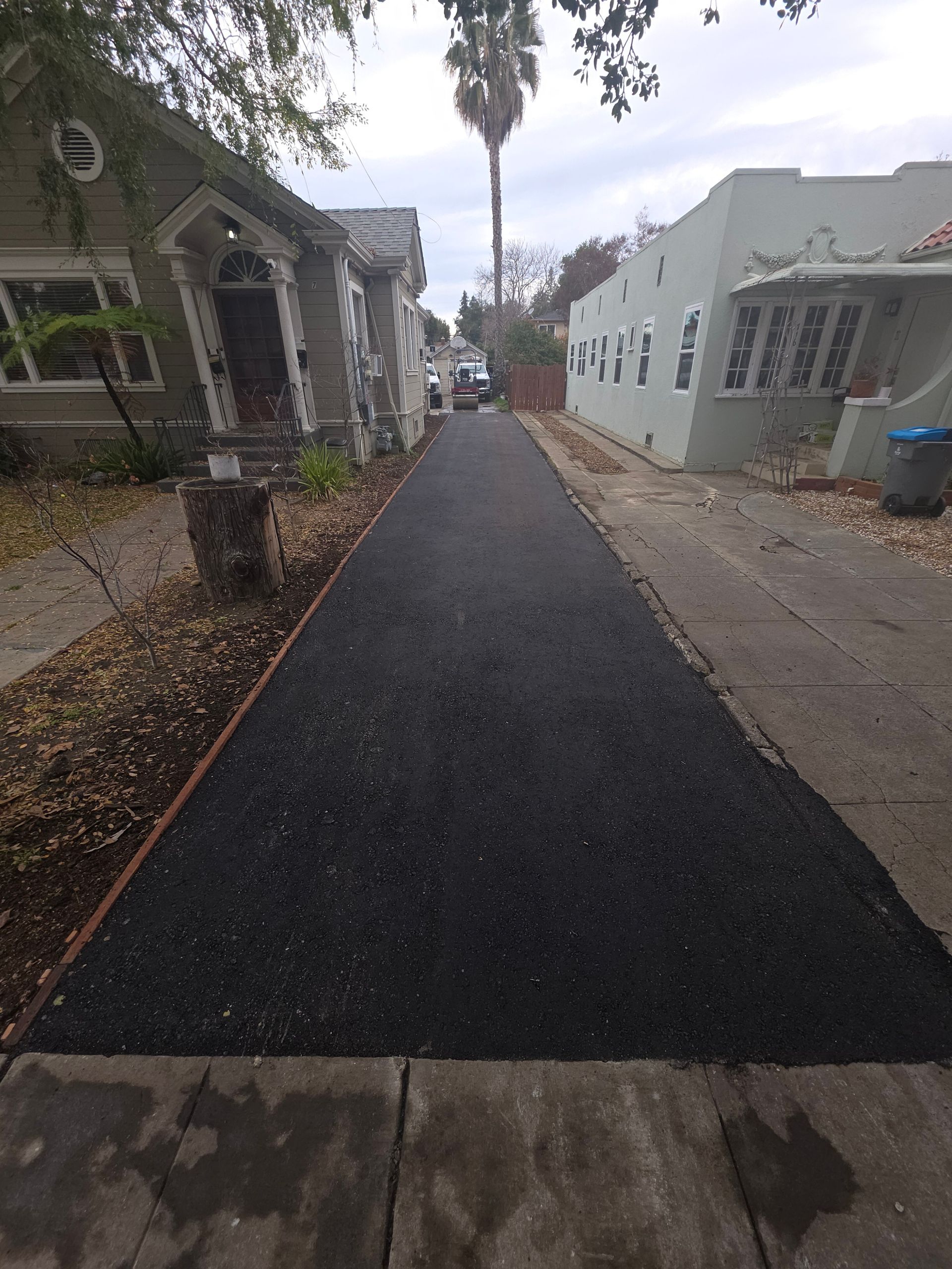 A newly paved asphalt driveway sits between two houses, extending from the sidewalk toward the back of the property.