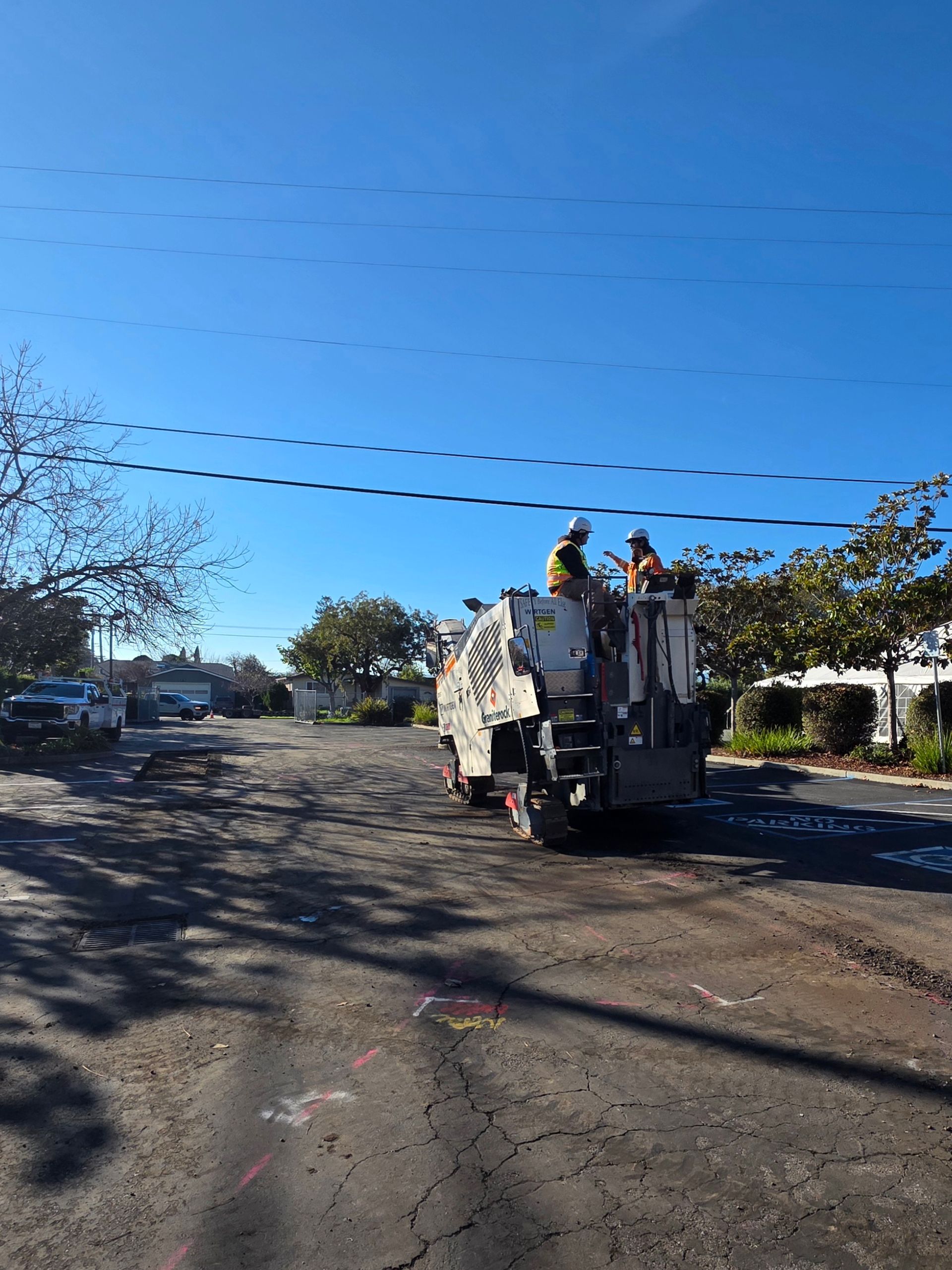 Two workers in safety gear operate a milling machine on a cracked, weathered asphalt street under a clear blue sky.