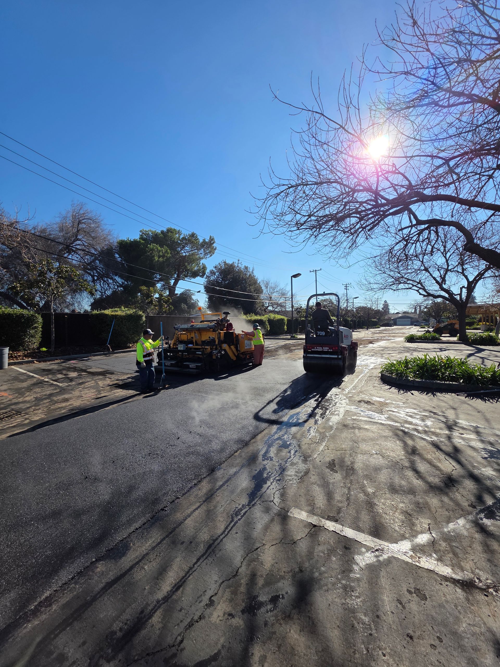 Road crew uses a paver and a roller to resurface a sunny suburban street.