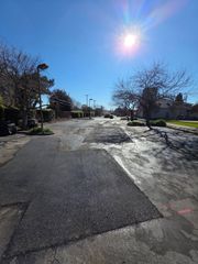 A residential street under a bright sun with several large, recently paved asphalt patches covering damaged road surface.