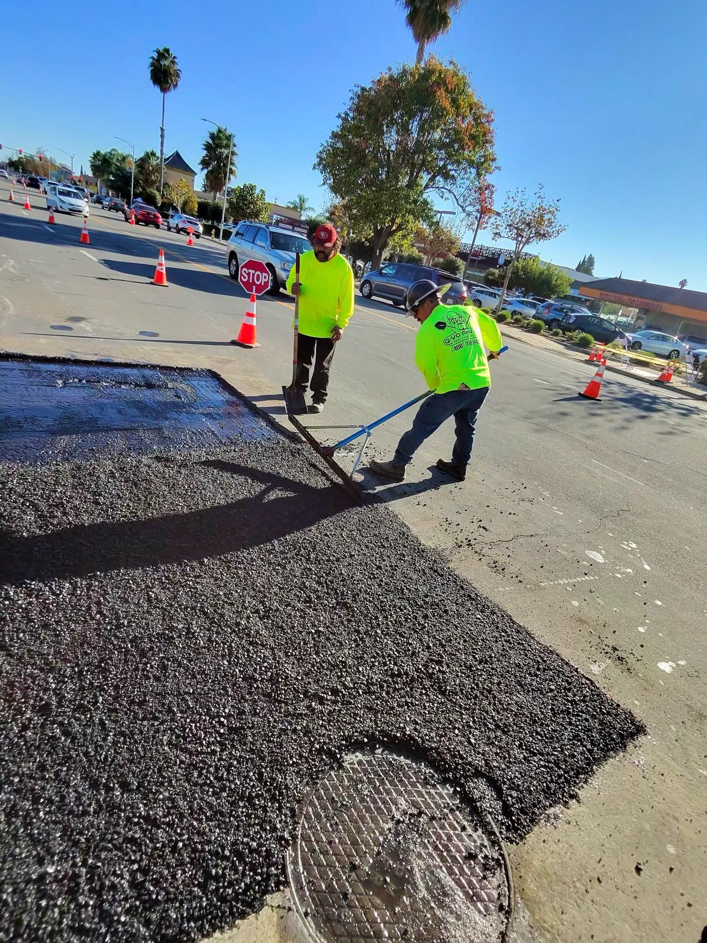 Two construction workers in neon yellow shirts spread fresh black asphalt on a road near a manhole cover.