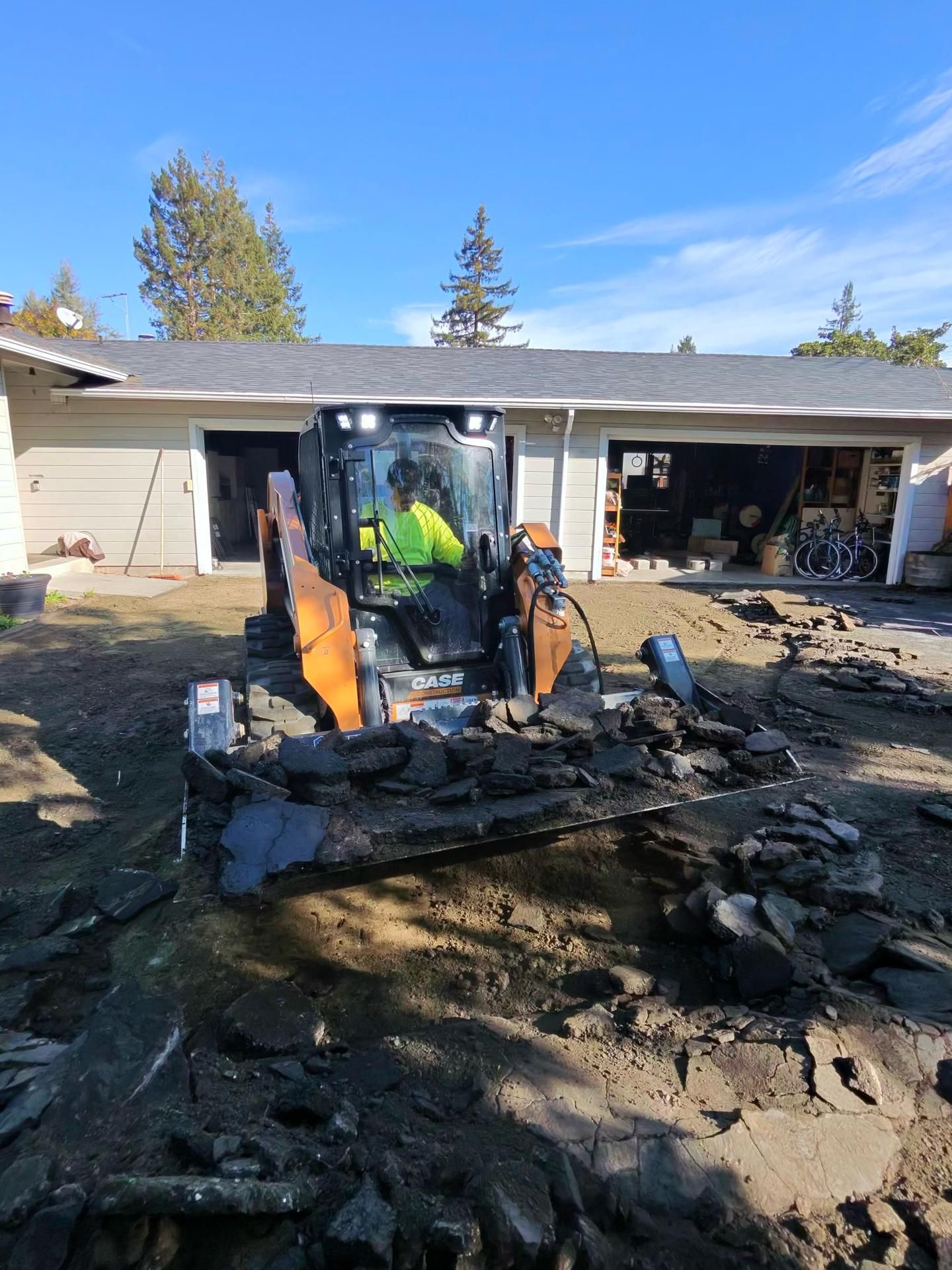 An orange skid-steer loader removes chunks of asphalt from a dirt driveway in front of a house.