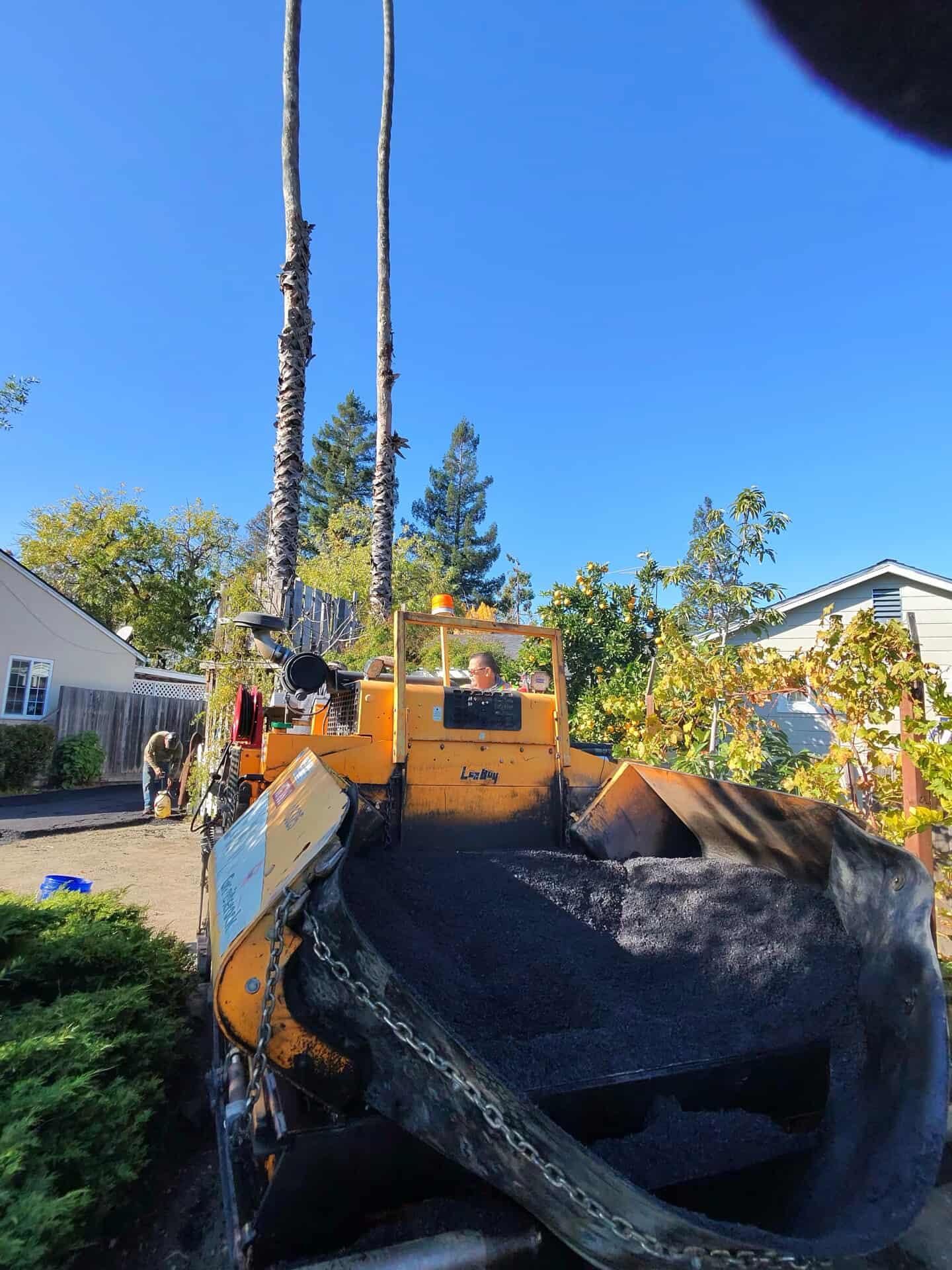 A yellow asphalt paver sits outdoors on a sunny day with a house and trees in the background.