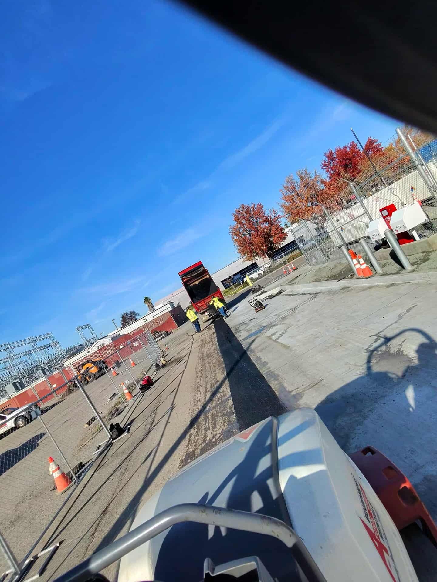 Construction workers in high-visibility vests work near a truck in a paved lot under a clear blue sky.