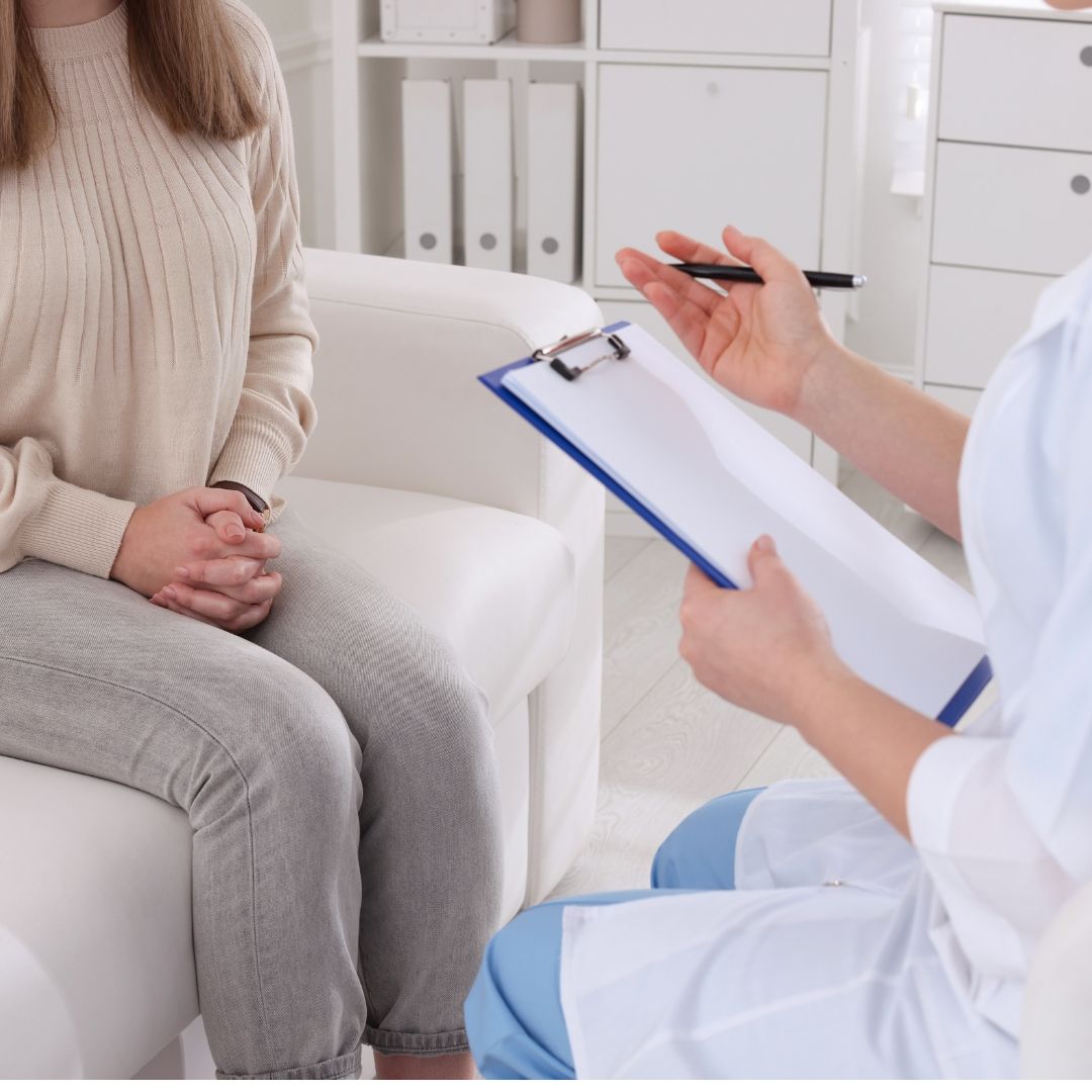 A woman in a sweater sits on a white couch while a medical professional, holding a clipboard and pen, speaks.