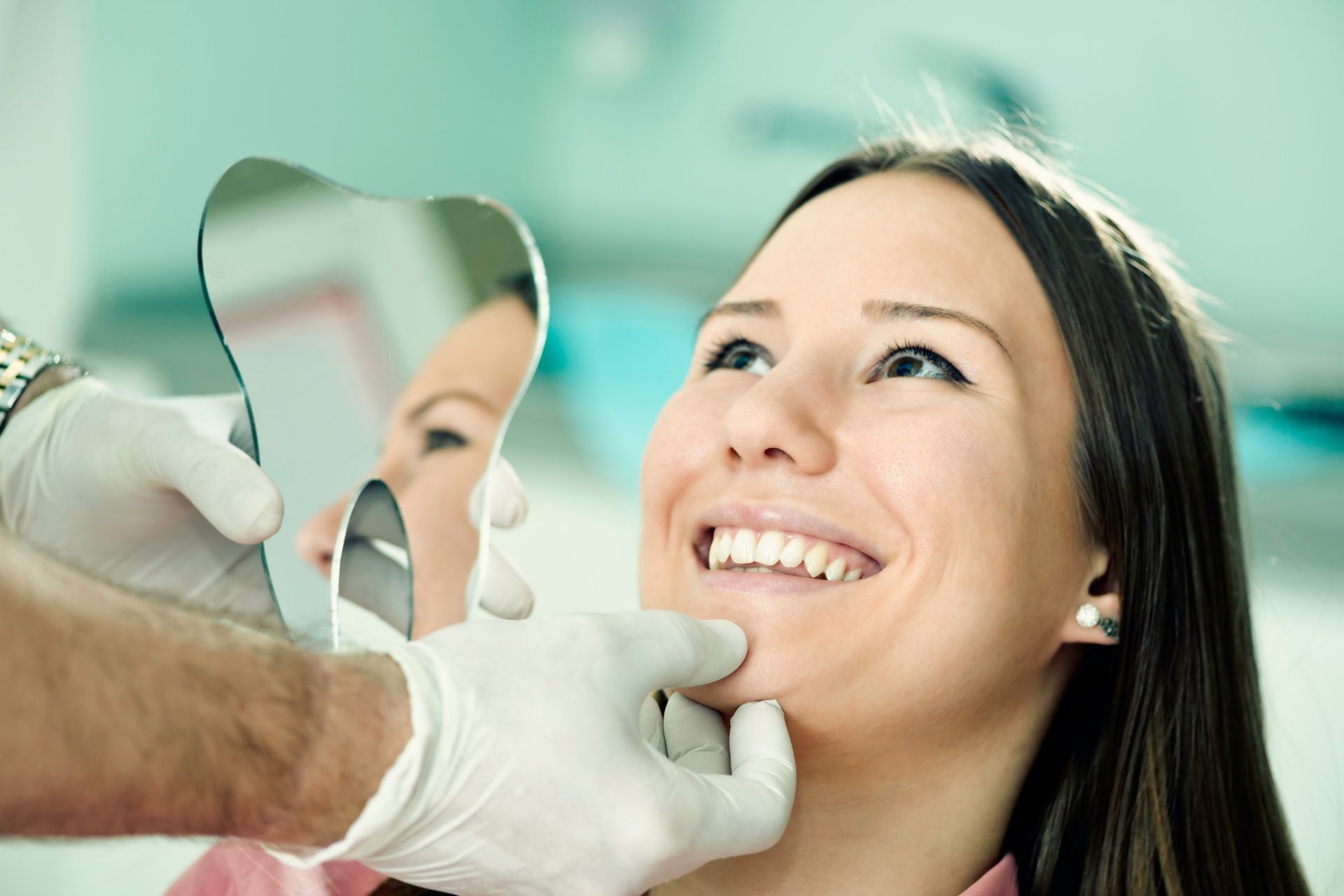 Dentist examining a smiling woman's teeth with a mirror in a bright dental office setting. Both are looking at the teeth, the dentist is wearing gloves.