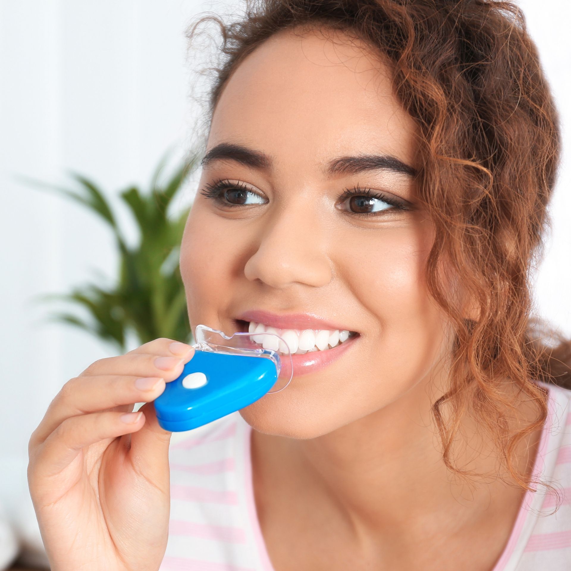 Woman holding a blue teeth-whitening device in her mouth, smiling. She has brown curly hair and is wearing a pink and white striped shirt.