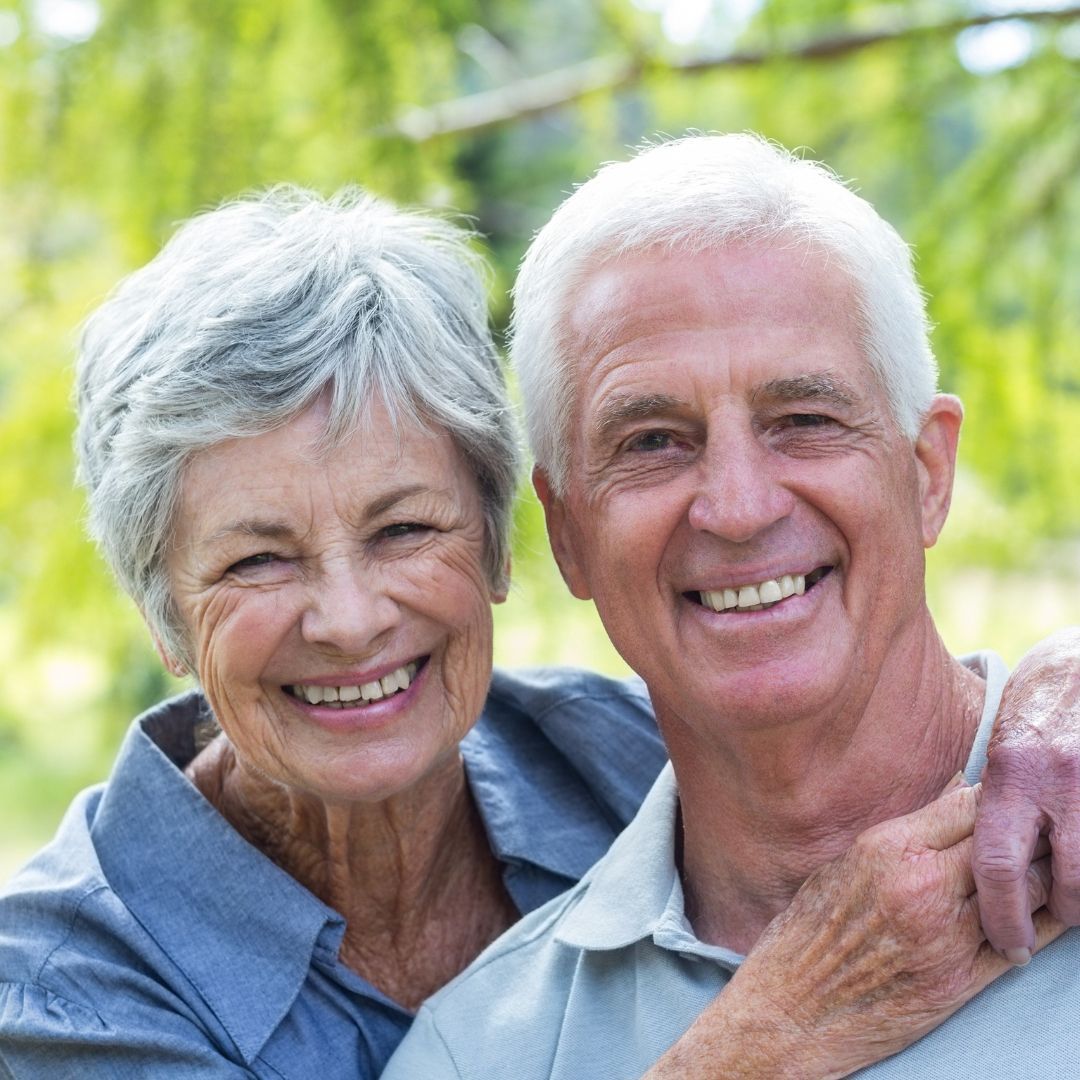 Smiling elderly couple embracing outdoors. The woman wears a blue shirt, and the man has his arm around her.