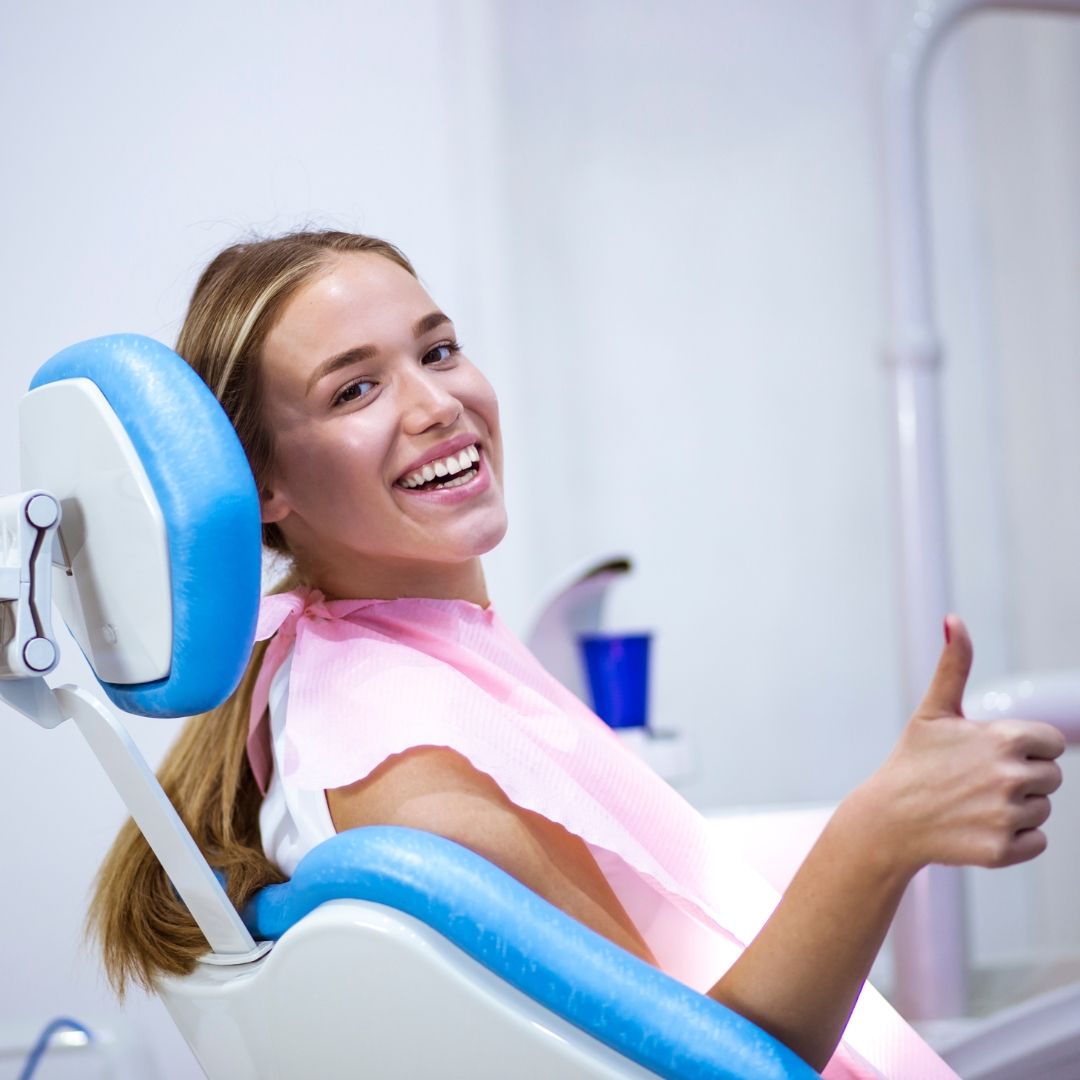 Woman in a dental chair gives a thumbs-up. She wears a pink bib, smiles, and has blonde hair.