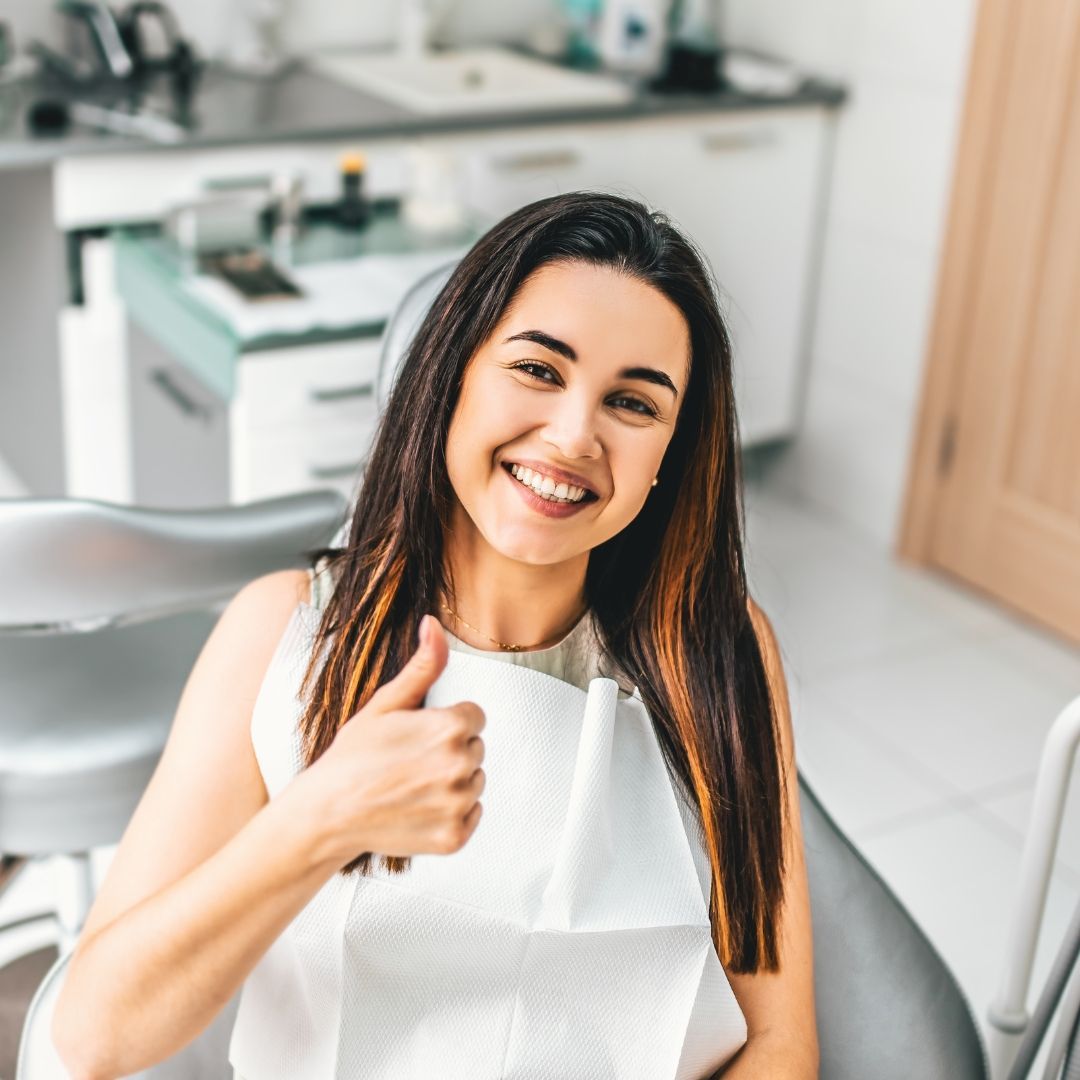 Woman smiling with a thumbs up in a dentist's chair, white bib, clinic setting.