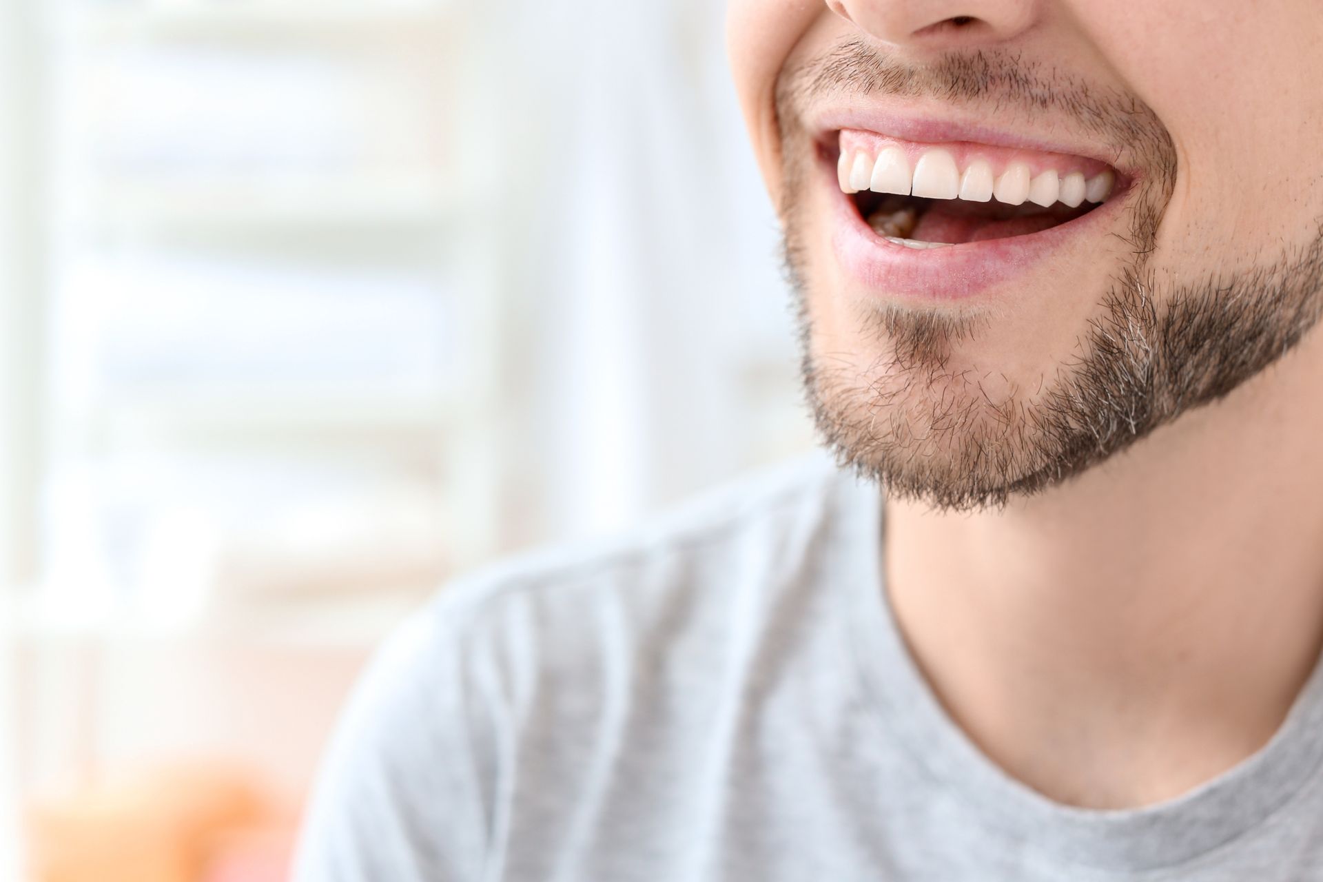 Man with a beard smiling widely, showing white teeth, wearing a gray shirt, indoors.