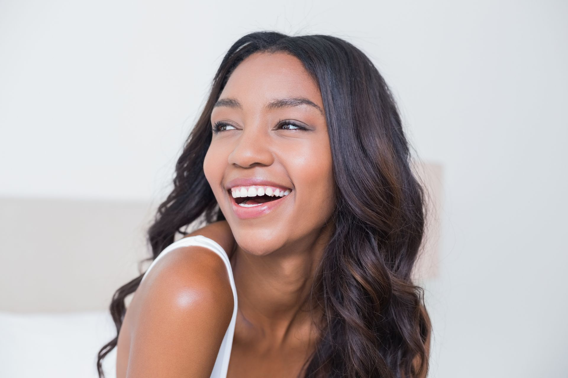 Smiling woman with long, dark wavy hair, looking off to the side, in a bright room.