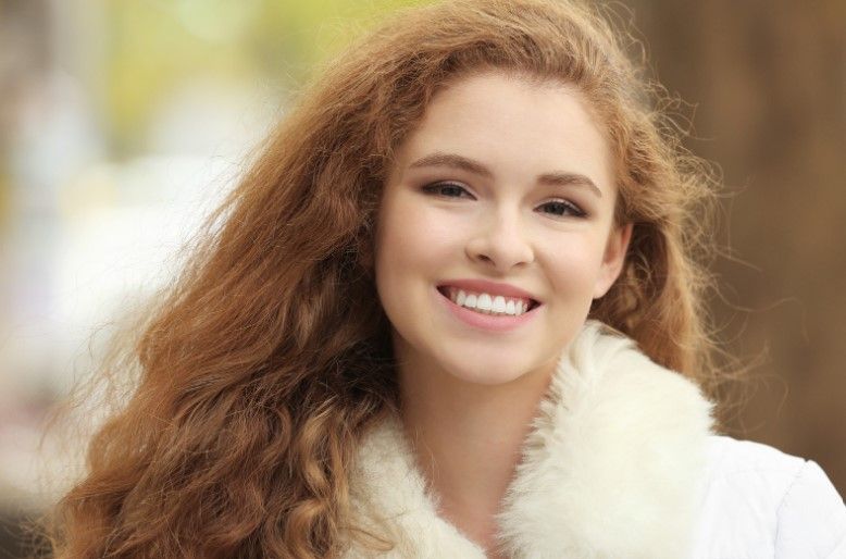 Woman with long, curly red hair smiles, wearing a white coat with a fur collar, outdoors.