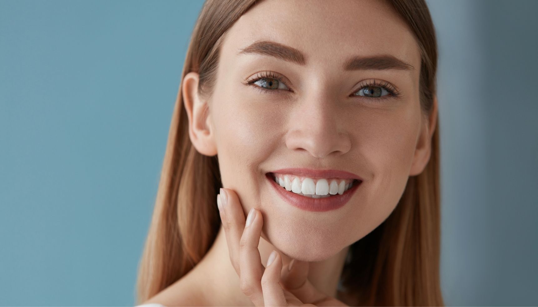 Smiling woman with fair skin and light brown hair, touching her cheek, against a blue backdrop.