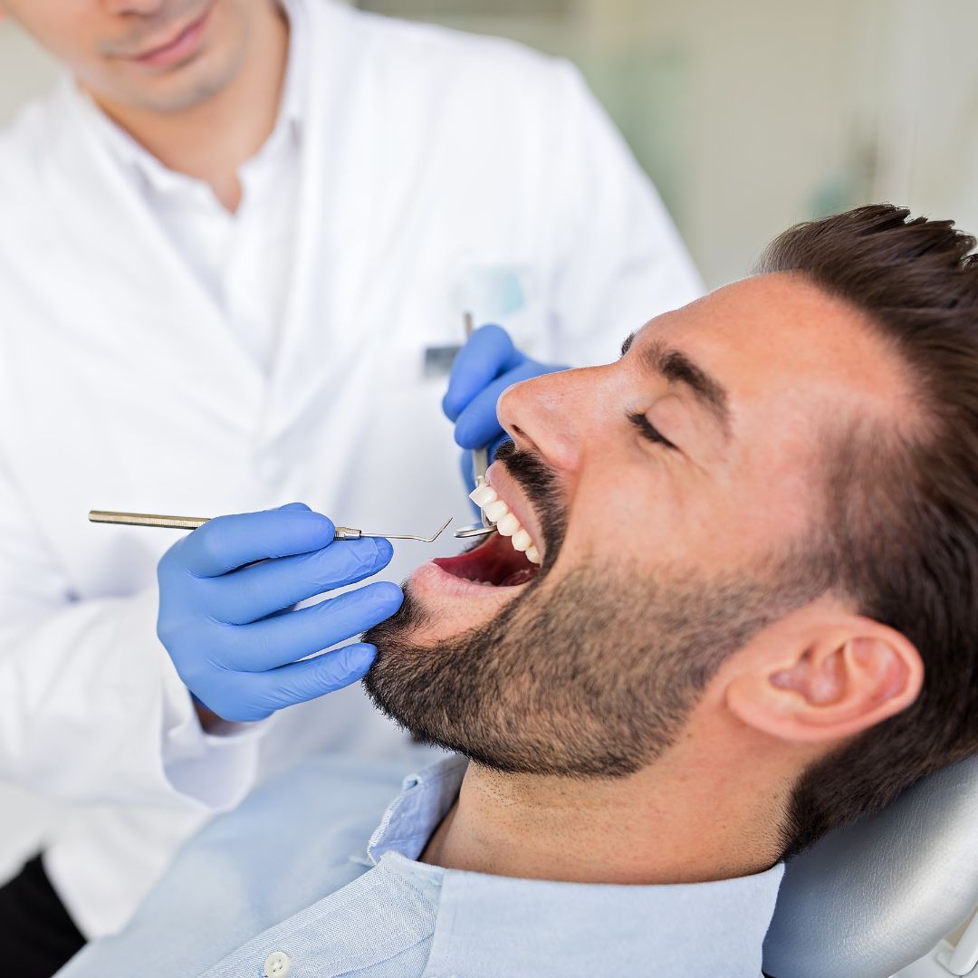 A dentist examining a man's teeth in a dental chair. The man has his mouth open, and the dentist is using instruments.