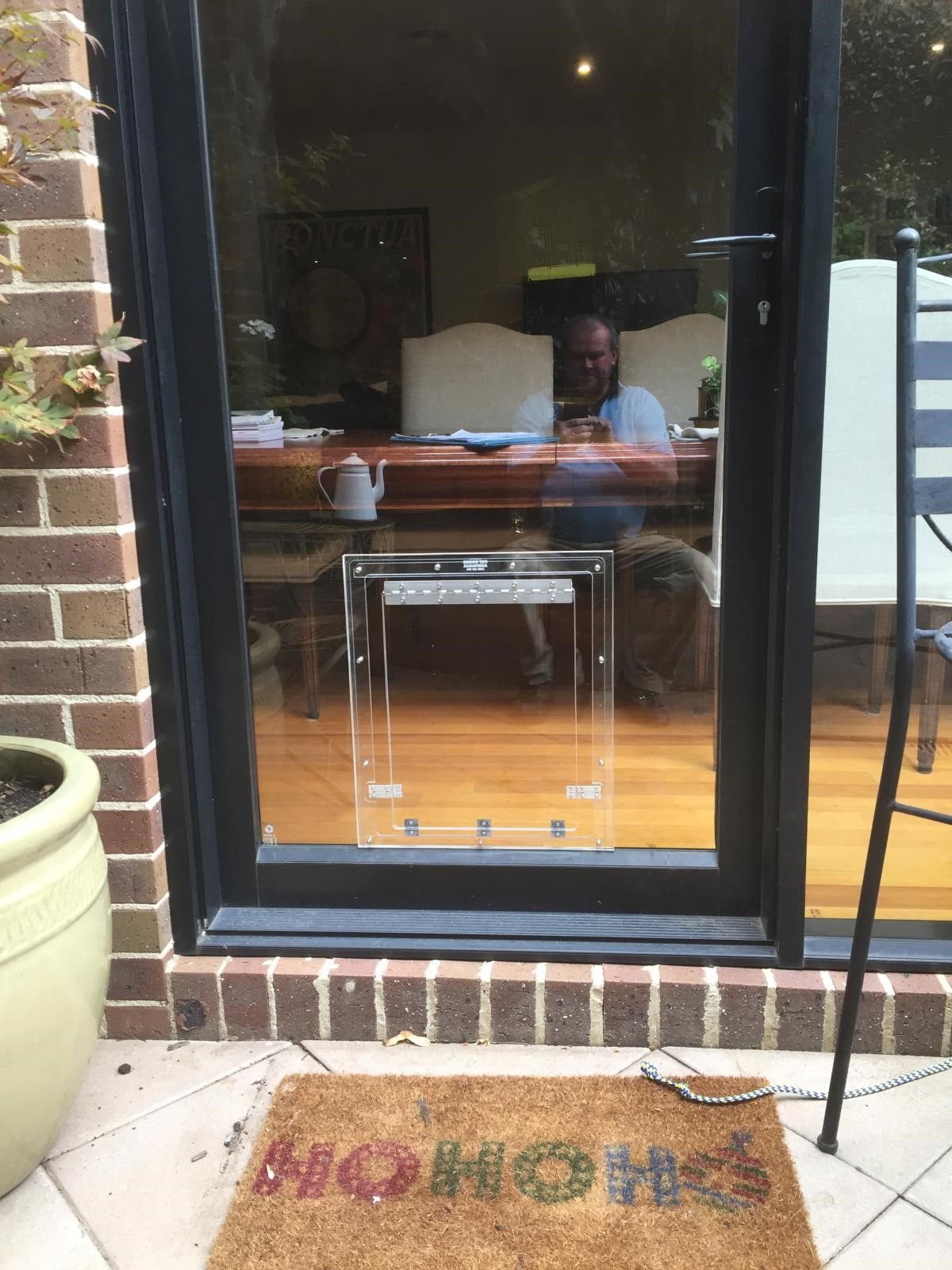Glass patio door reflecting a person inside; “home” doormat on brick patio beside a large planter.