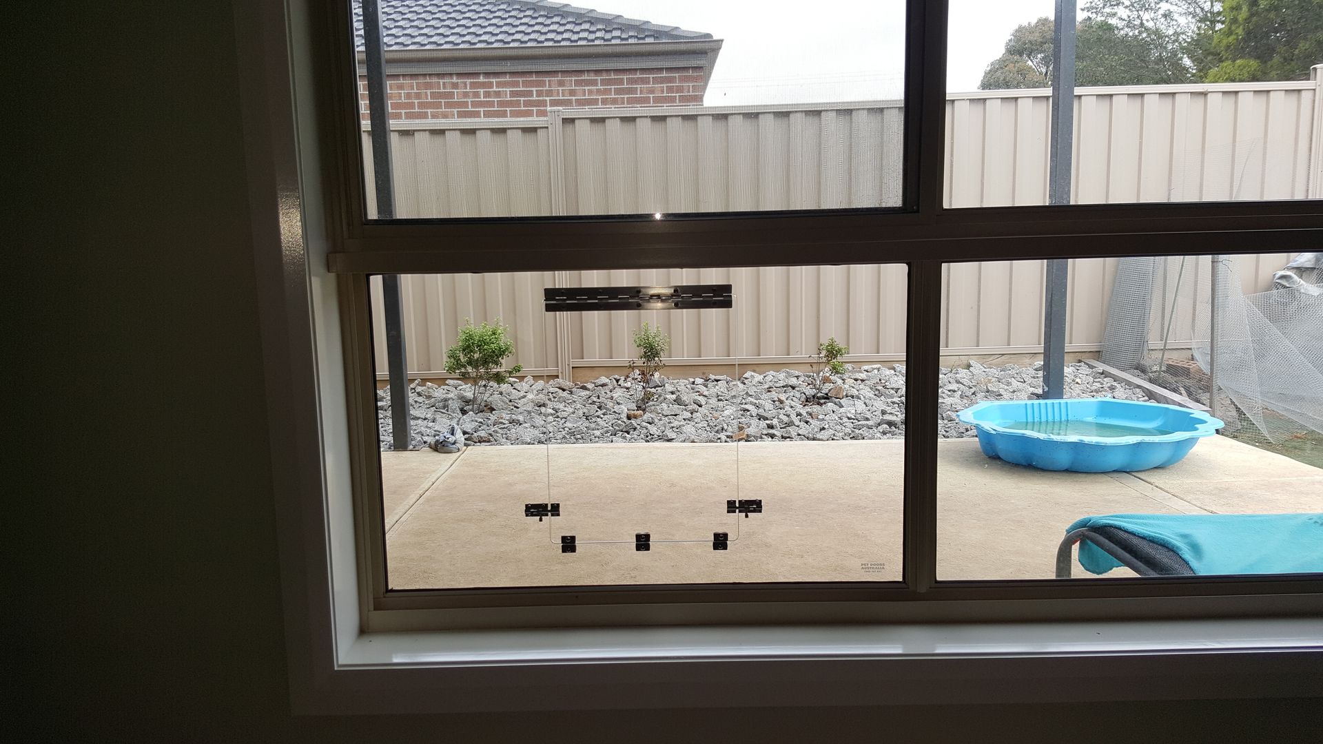 View through a window of a backyard with a blue kiddie pool, gravel, and a fence.