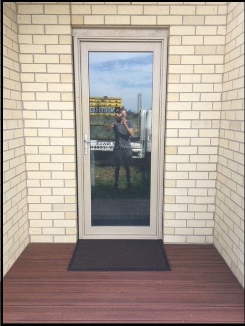 Glass door in beige brick wall with black doormat and reflected person outside.