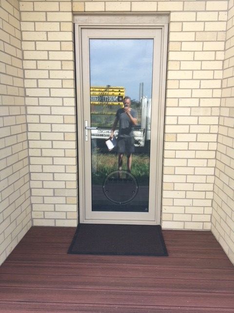 Glass door in beige brick wall, with a person reflected on the dark mat-covered entrance.