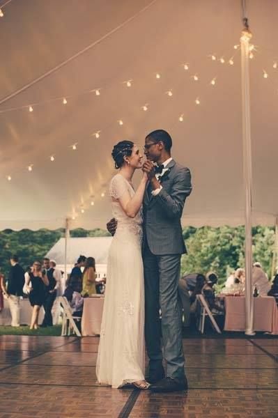 Bride and groom dance under a tent with string lights; guests seated at tables.