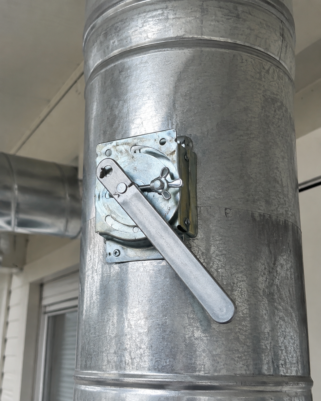 Metal duct with a gray lever handle mounted on a wall in an indoor industrial space