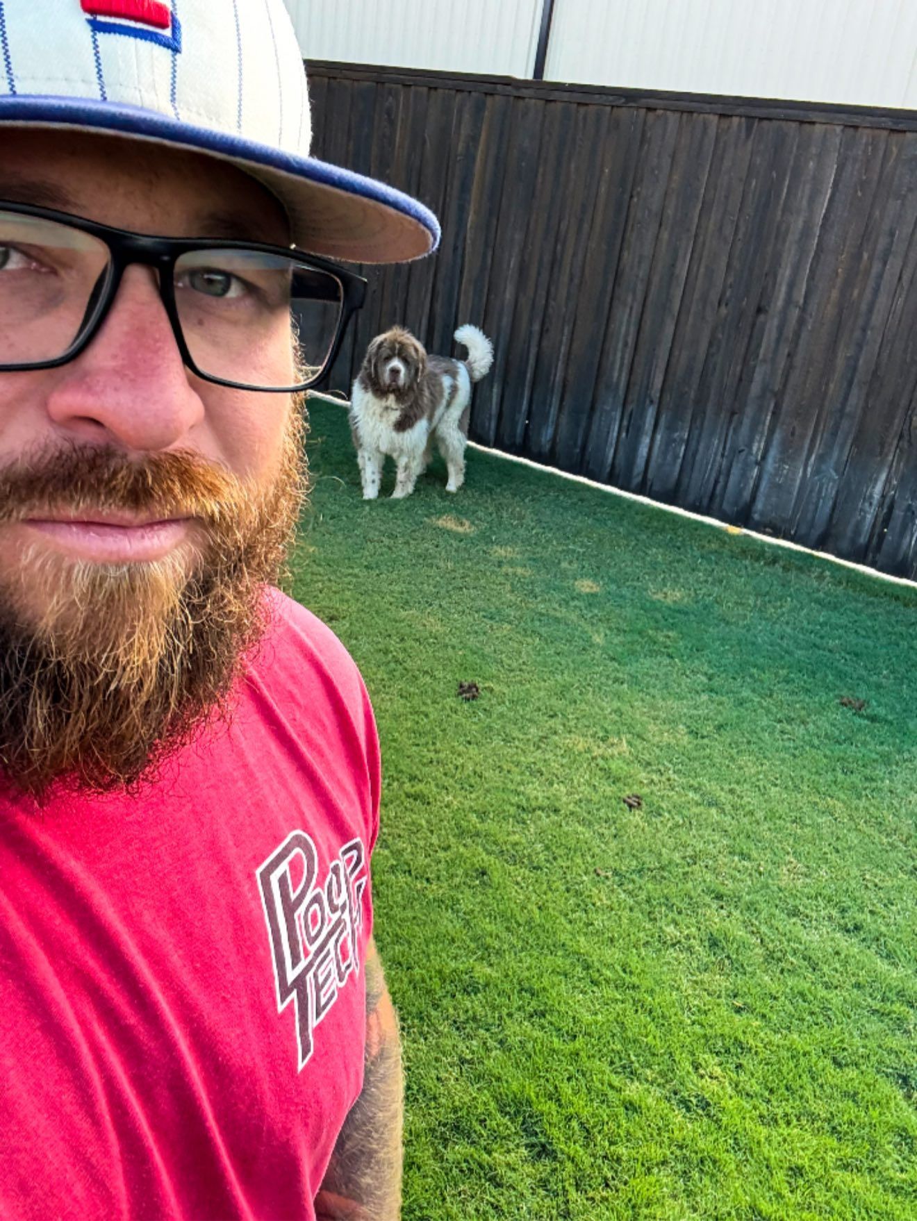 Man in glasses and red shirt taking a selfie in a backyard with a small dog near a black fence.
