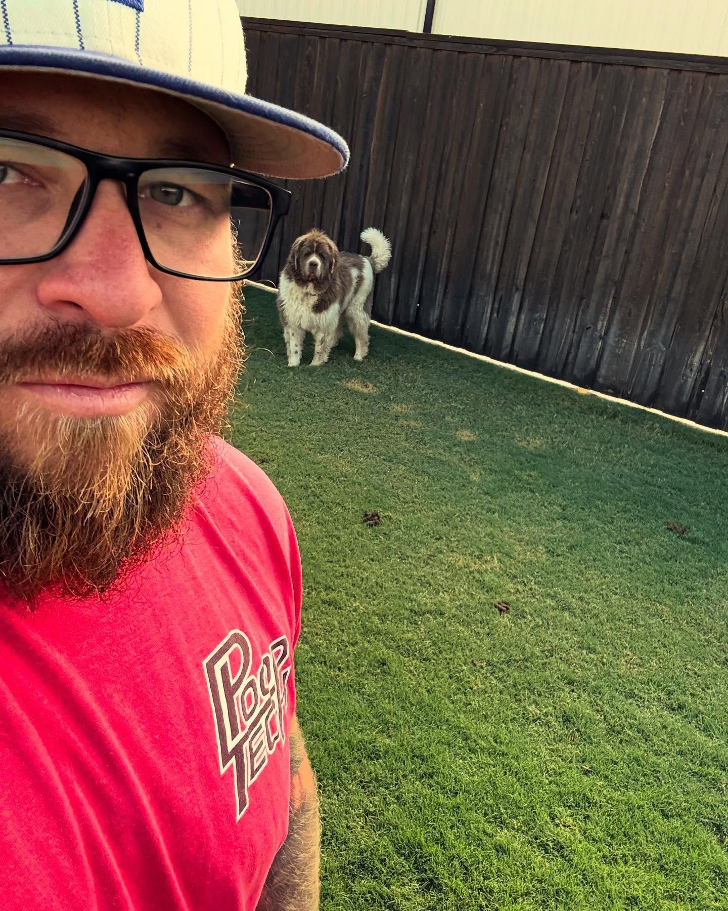 Selfie of a man in a red shirt and hat, with a dog on grass in a fenced yard behind him