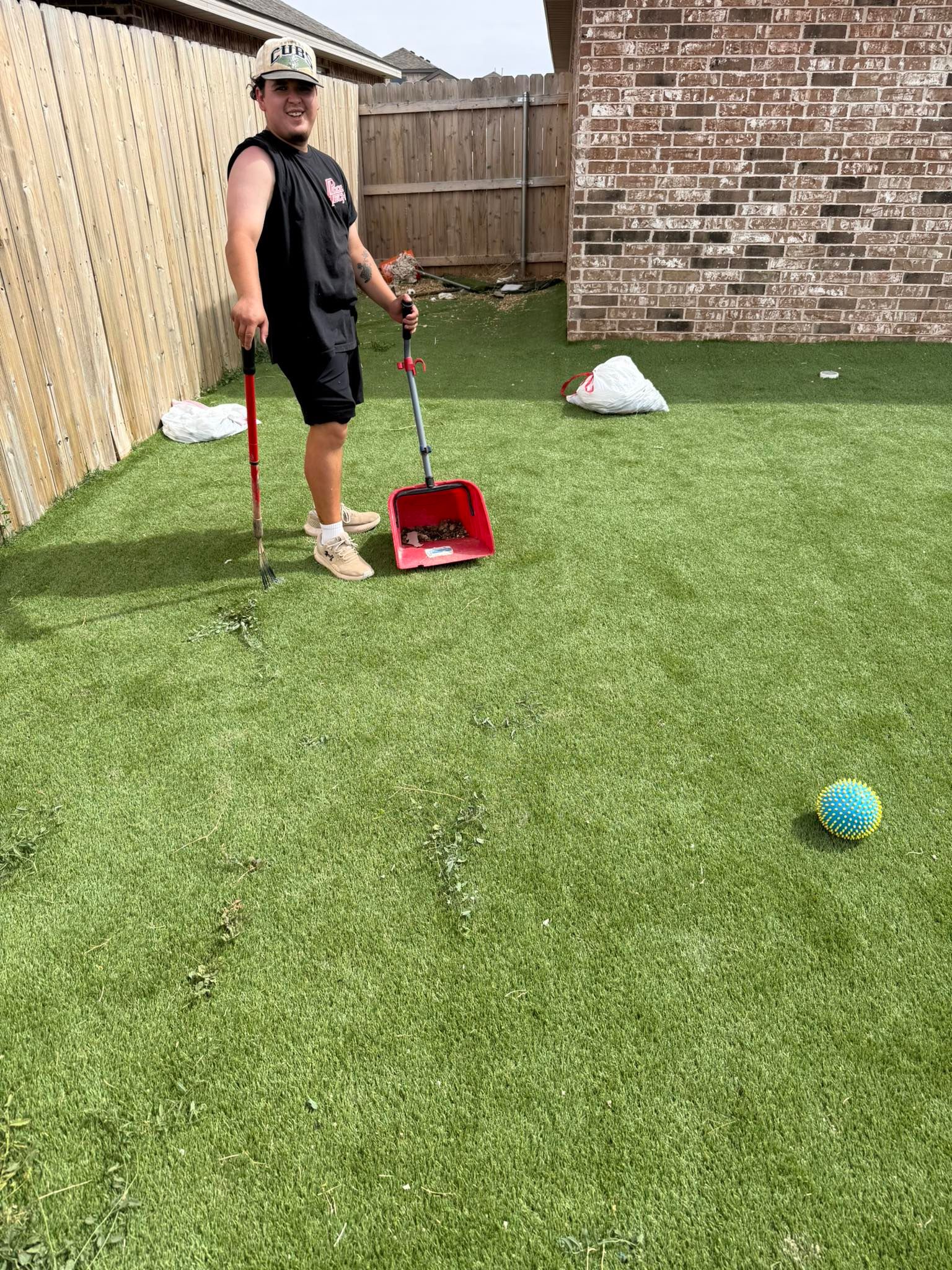 Person standing on artificial turf holding a red leaf blower beside scattered white bags and a blue ball.
